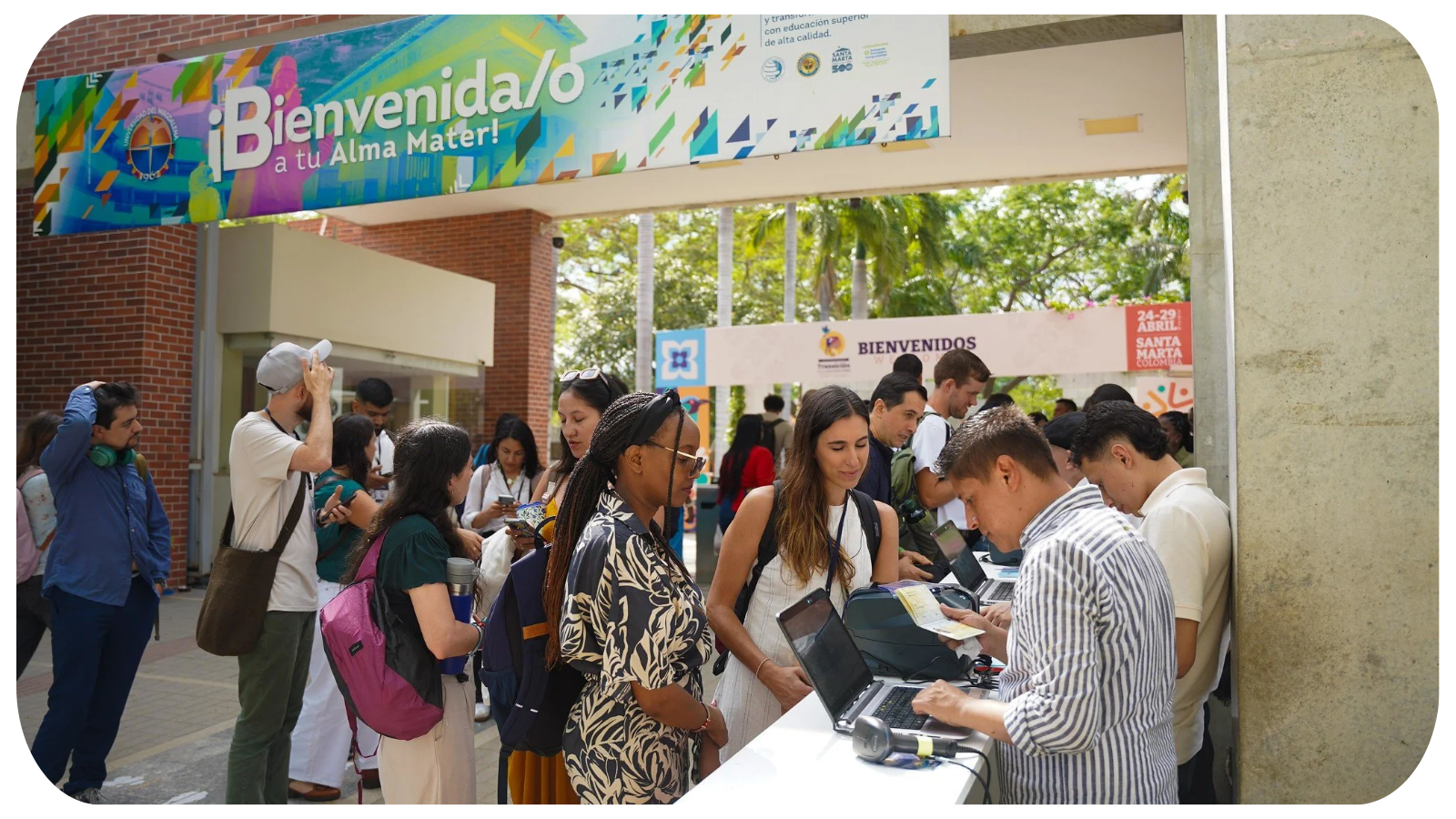 People waiting in line at an event registration booth under a colorful banner that says "Bienvenida/o a tu Alma Mater!" with greenery and trees in the background.