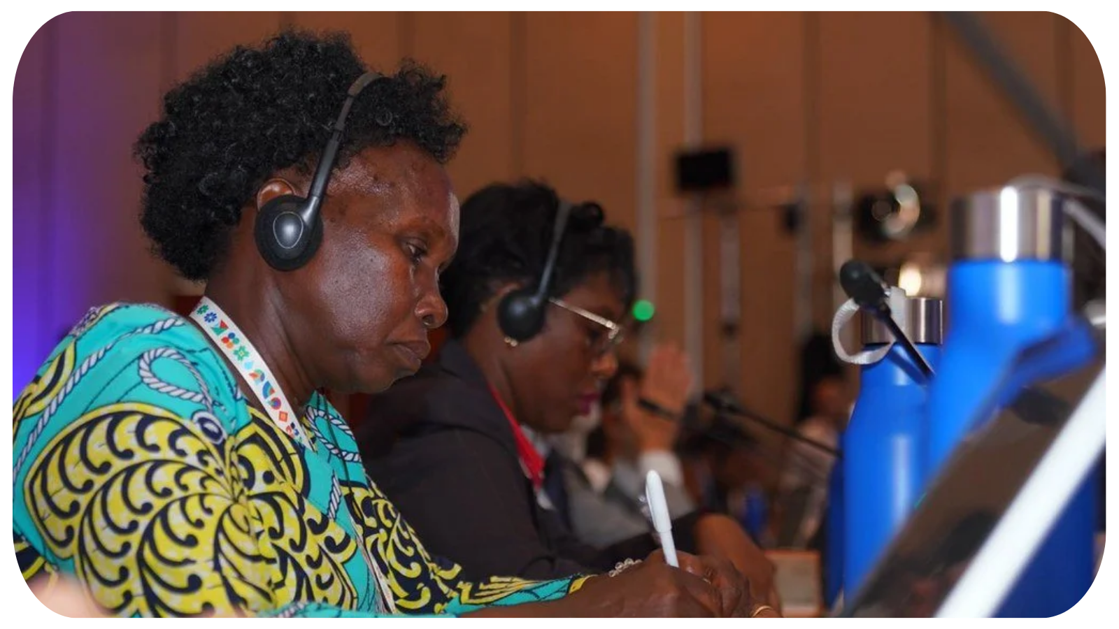 Two women sitting at a conference table with headsets, taking notes, and looking at laptops during a professional event.