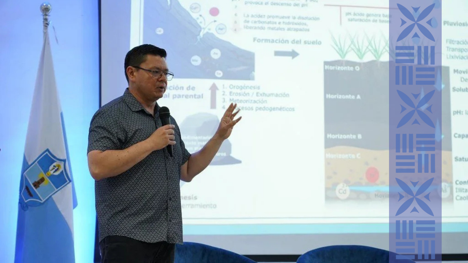 A man wearing glasses and a patterned shirt is giving a presentation while holding a microphone. Behind him, there is a large screen with slides and a blue and white flag with a coat of arms.
