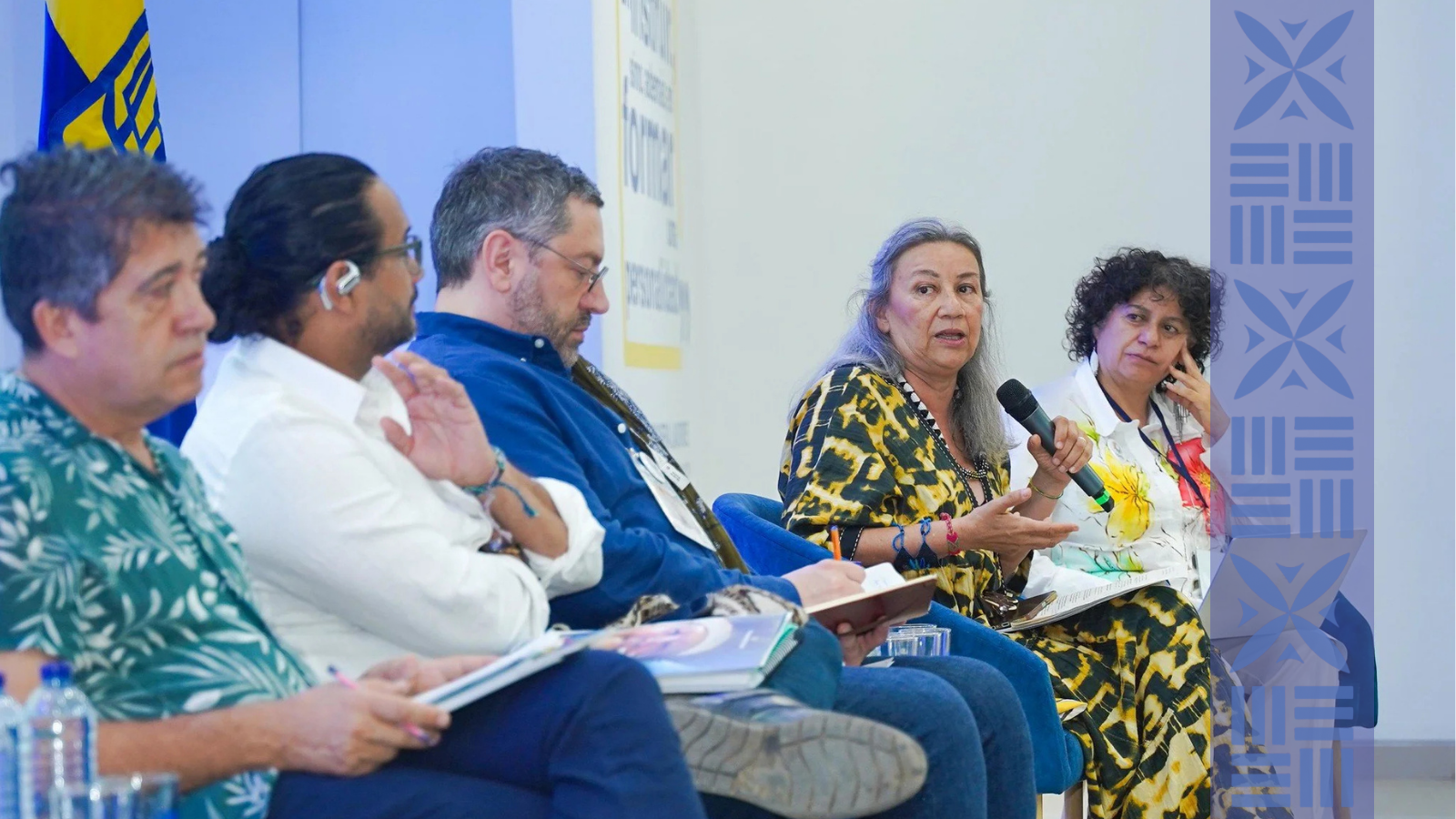 A group of people sitting on a panel at a conference, one woman speaking into a microphone while others listen, with a flag in the background.