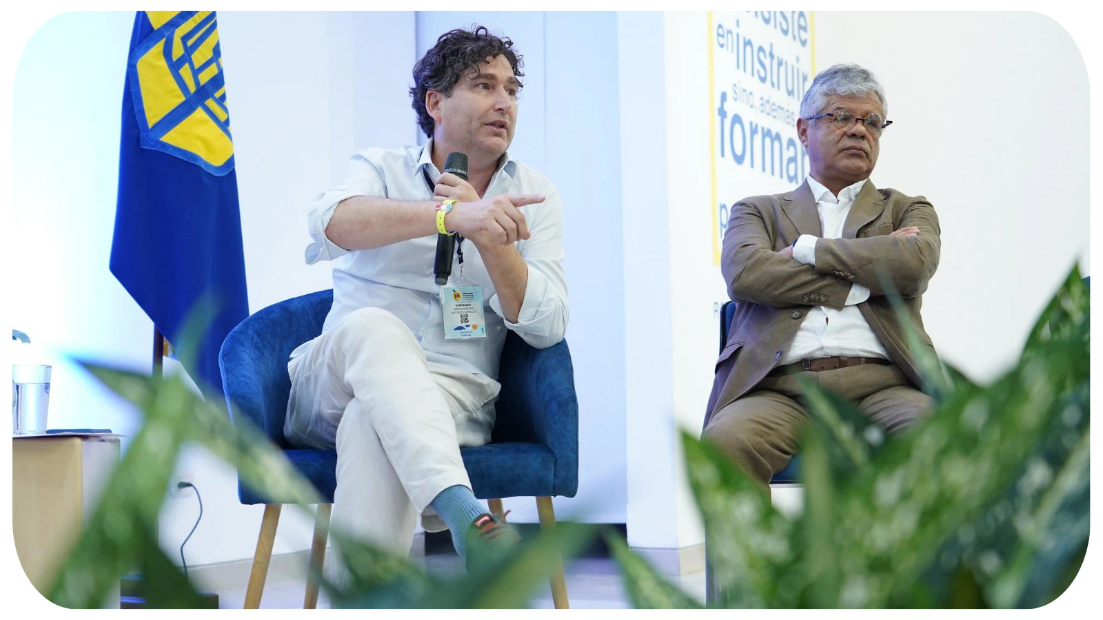 Two men sitting on stage in conversation at a conference, with a flag in the background and a plant in the foreground.