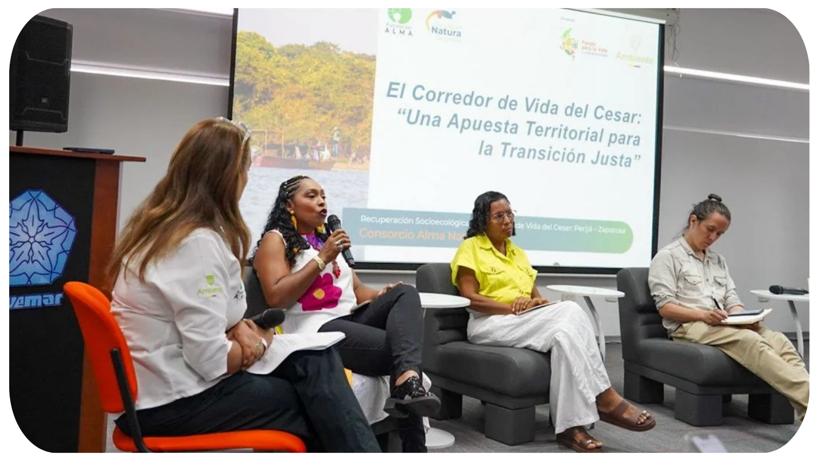Four women seated on a stage, two holding microphones and taking notes, in front of a large presentation screen displaying a title in Spanish about the Cesar River corridor and ecological recovery.