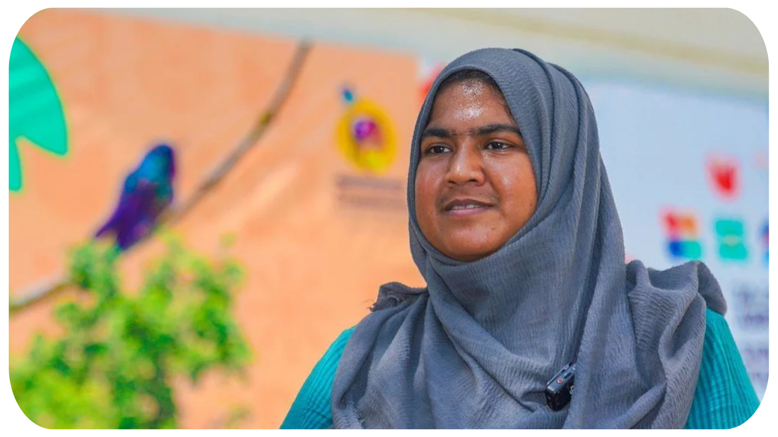 A woman wearing a blue hijab and teal outfit standing in front of a colorful background, with a blurred bird and plants in the foreground.