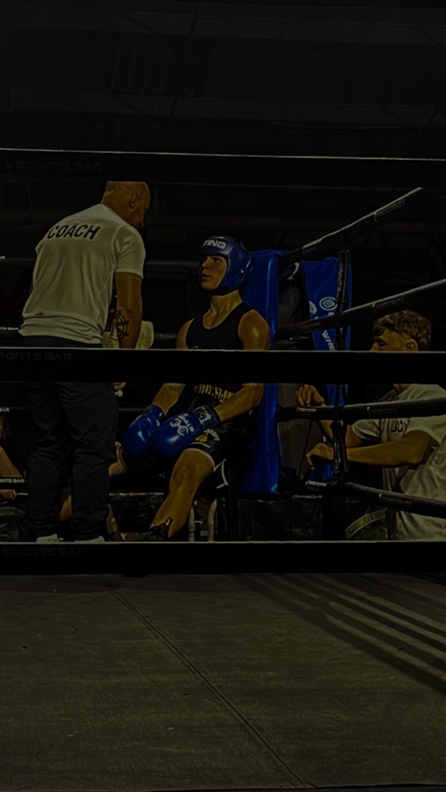 A boxer in blue shorts and headgear sitting in a corner of a boxing ring, talking with a coach who is standing next to him, while another person is sitting in the corner holding a water bottle.