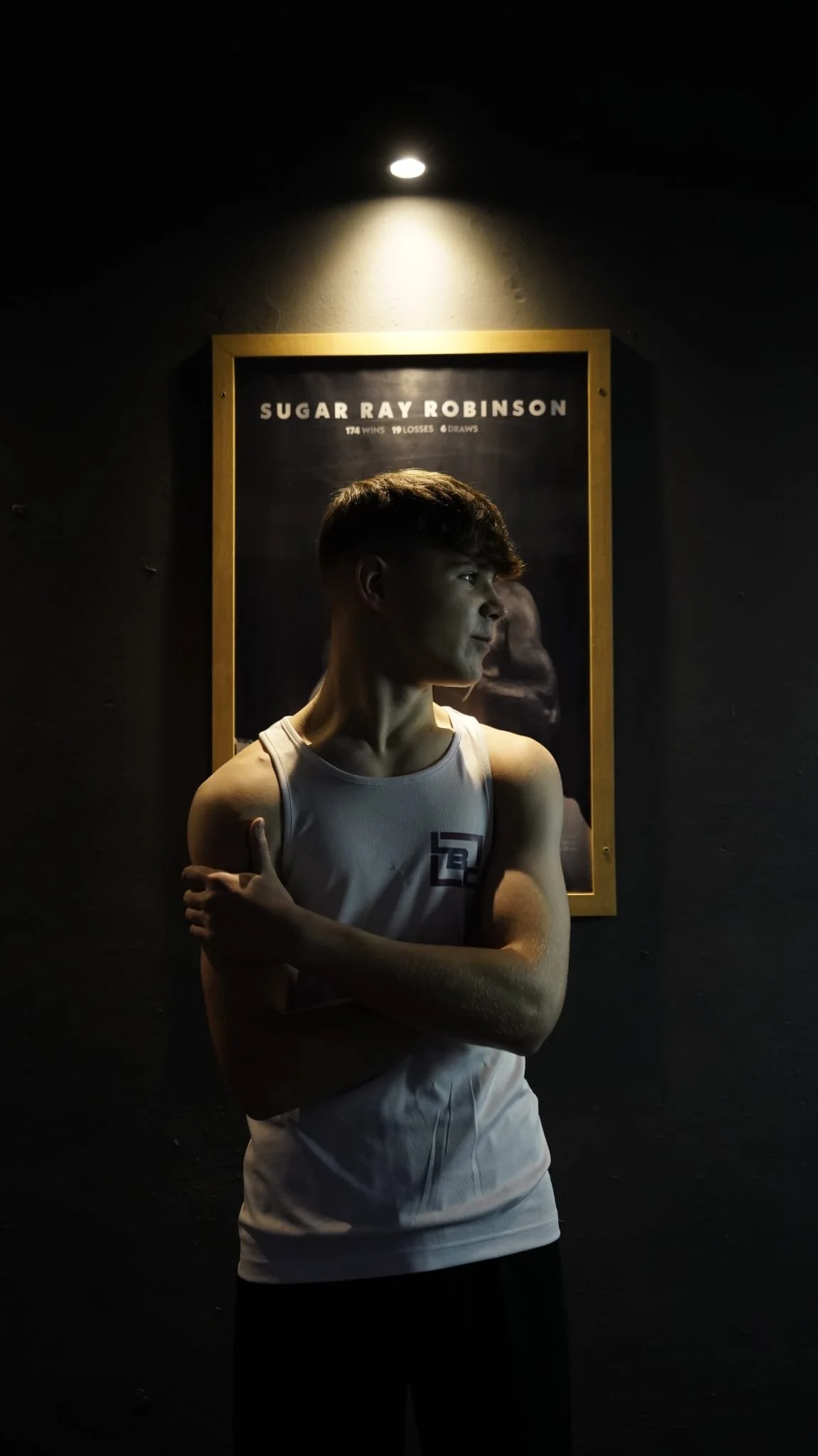 A young man with short dark hair wearing a white sleeveless athletic shirt standing in front of a black wall. Above him is a framed poster of Sugar Ray Robinson with their fight record. He is crossing his arms, with one hand on his shoulder.