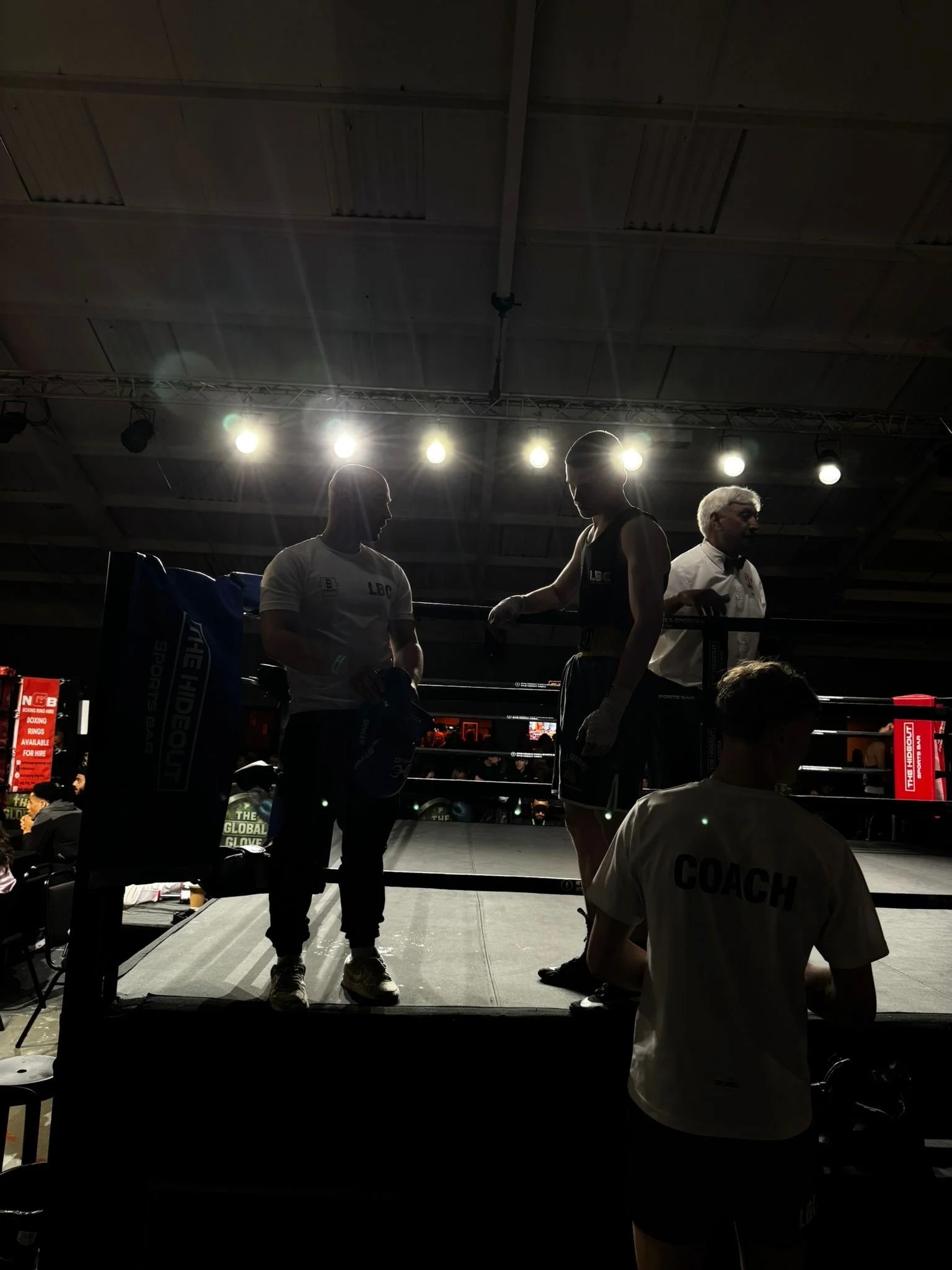 Boxing ring with two fighters and a referee under bright lights, with coaches and spectators around.
