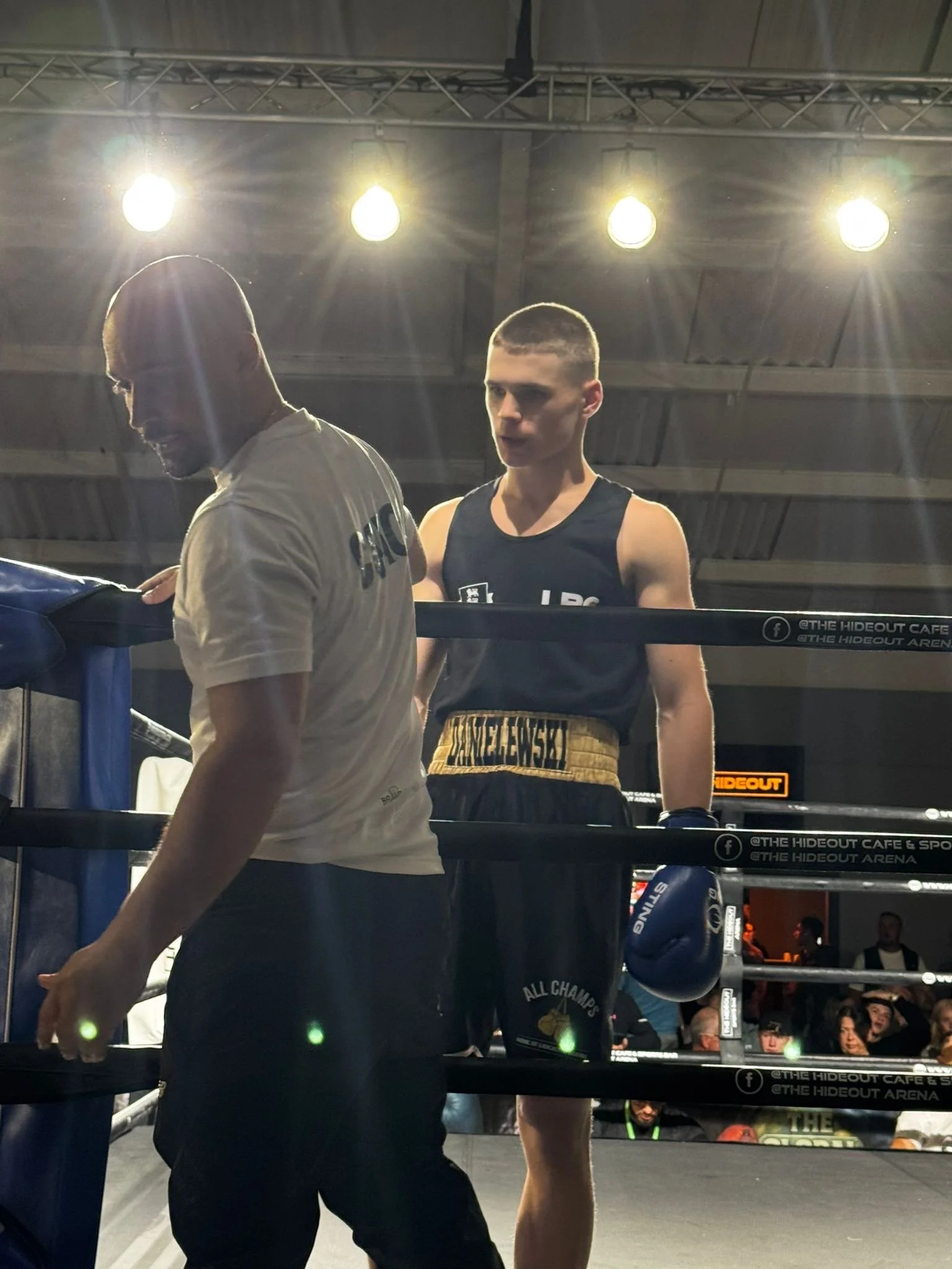 A boxing match in progress inside a ring, with a boxer wearing black shorts and a black tank top standing, and another person, possibly a coach or cornerman, in the foreground. Bright overhead lights illuminate the scene, with spectators visible in the background.