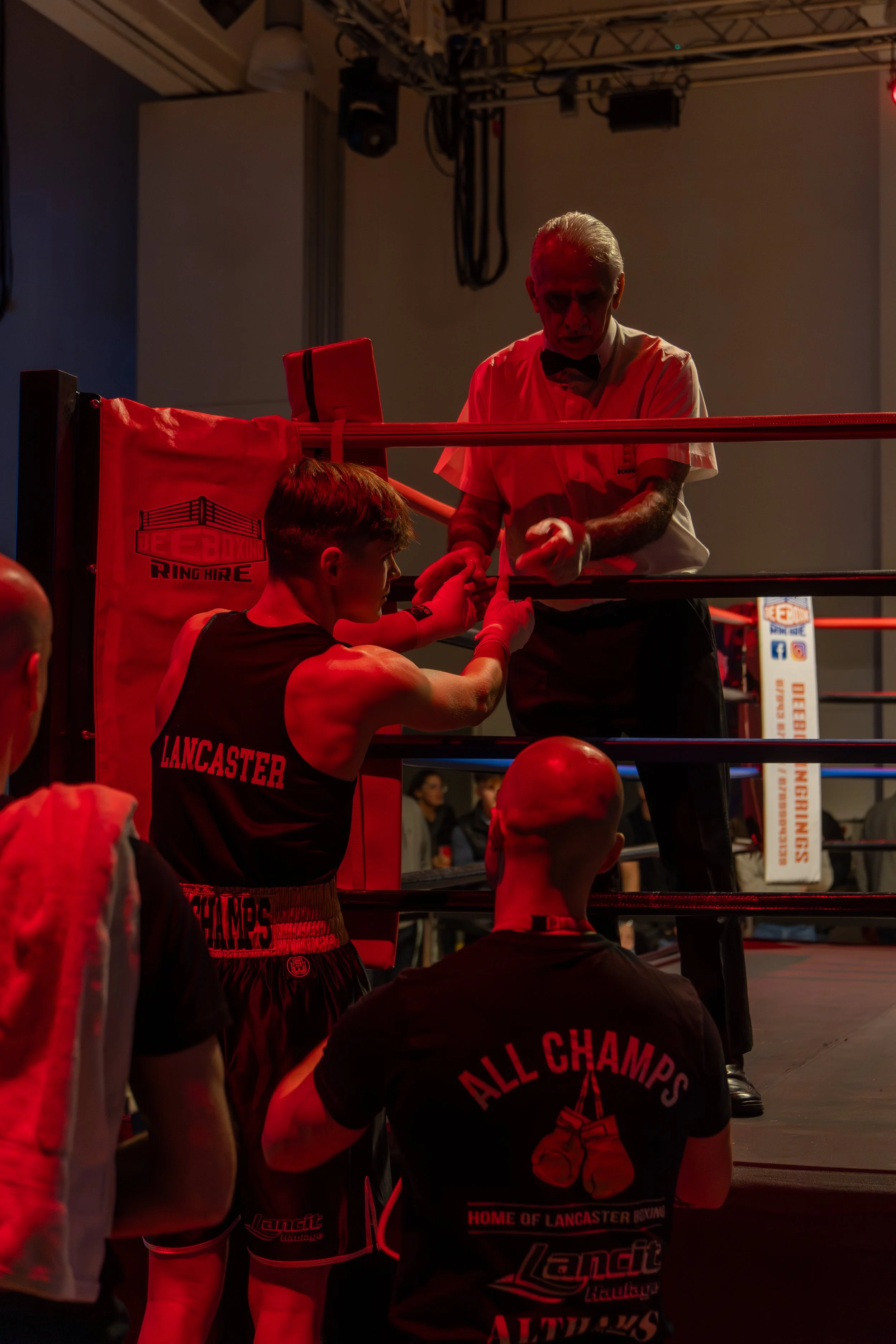 Boxer sitting inside a boxing ring receiving instructions from his coach, with other fighters and spectators around, illuminated by red lighting at a boxing gym.