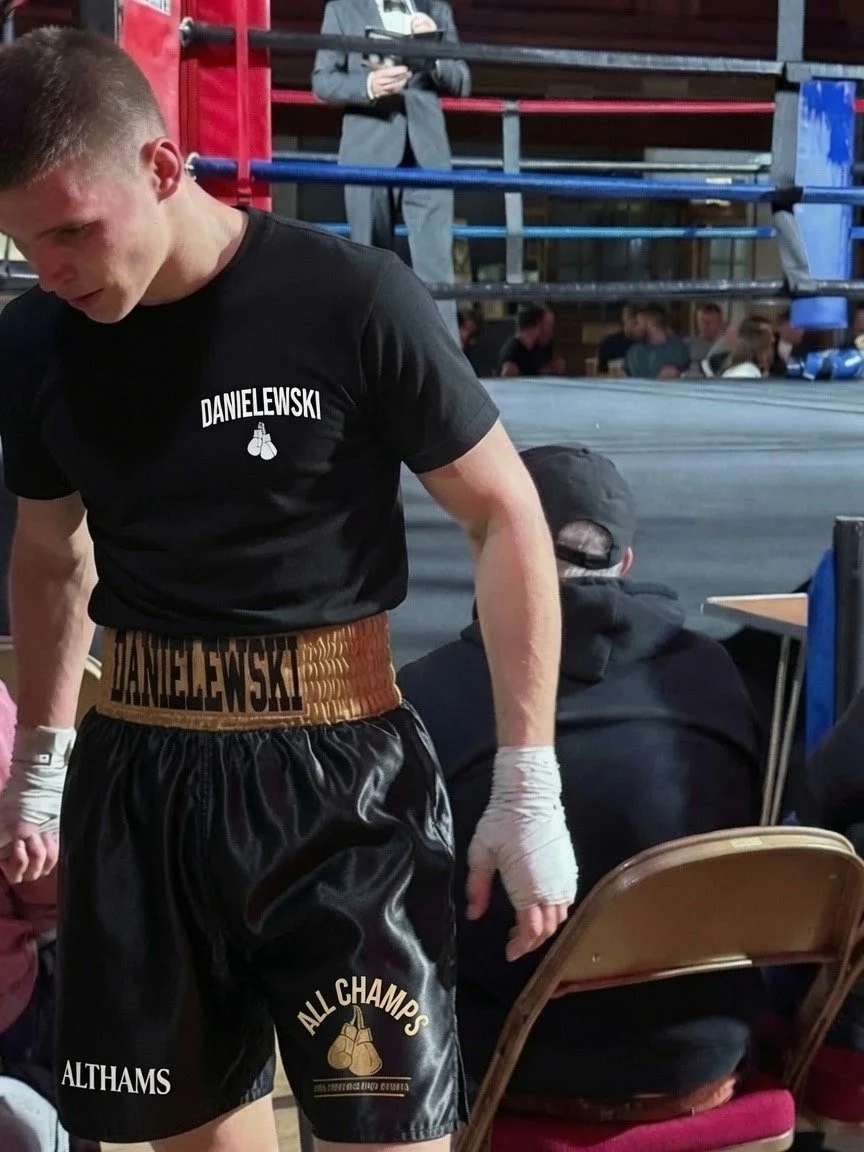Young male boxer in boxing shorts and gloves, standing in a boxing gym, surrounded by people and boxing ring.