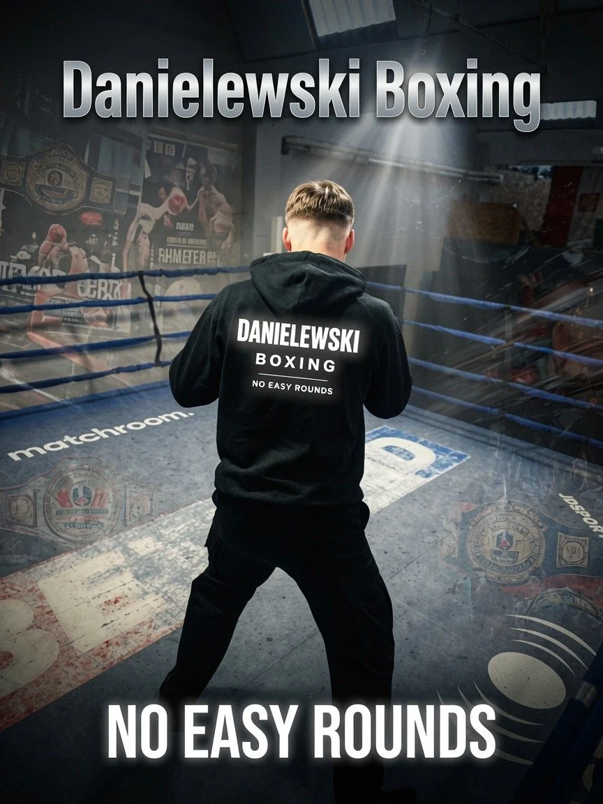 A boxer standing inside a boxing ring with his back facing the camera, wearing a black hoodie that says 'DANELEWSKI BOXING NO EASY ROUNDS'. The background shows boxing posters and championship belts, with the words 'Danelewski Boxing' and 'No Easy Rounds' overlaid on the image.
