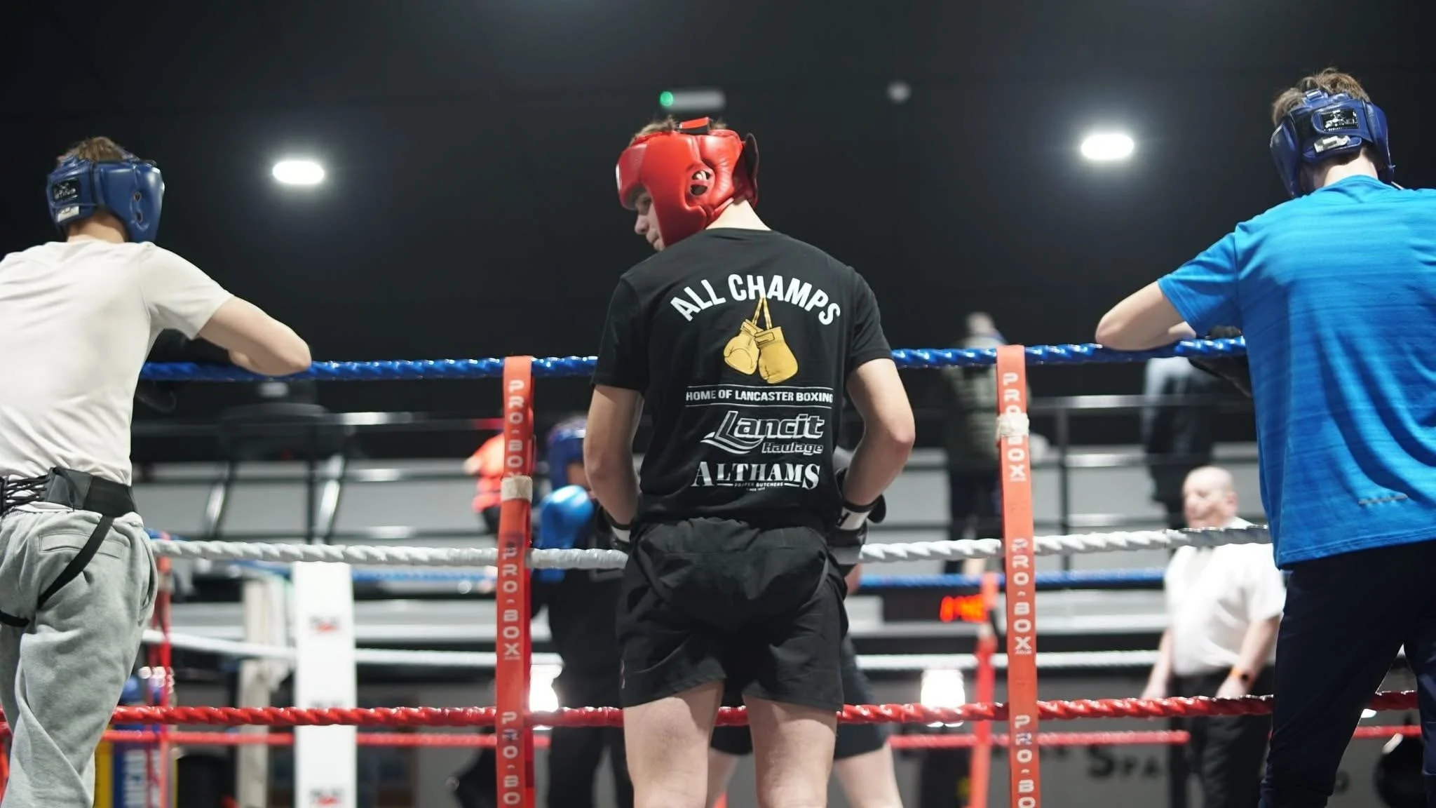 Boxers in a boxing ring, wearing headgear and shorts, preparing for a match, with one wearing a black shirt that says 'All Champs'.