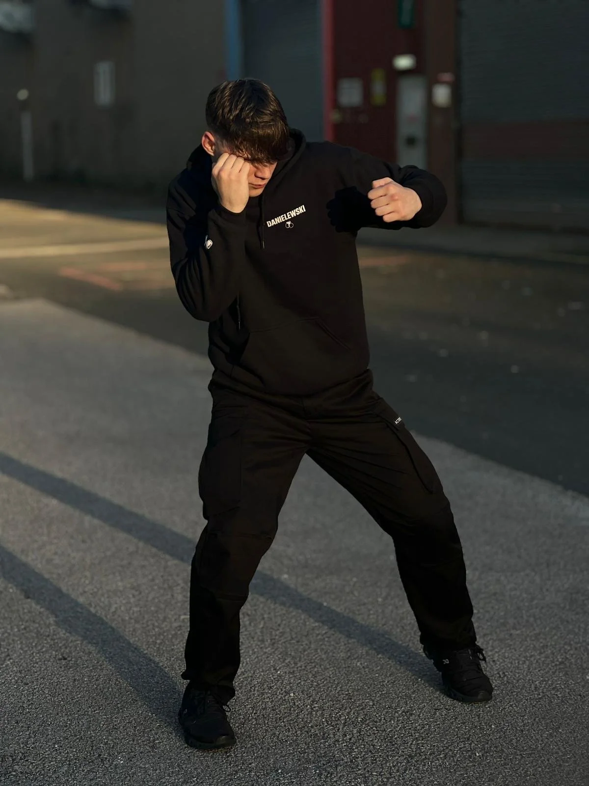 A young man wearing all black, including a hoodie with 'DANIELEWSKI' on it, standing on a city street at sunset, with one hand near his face and the other clenched into a fist.
