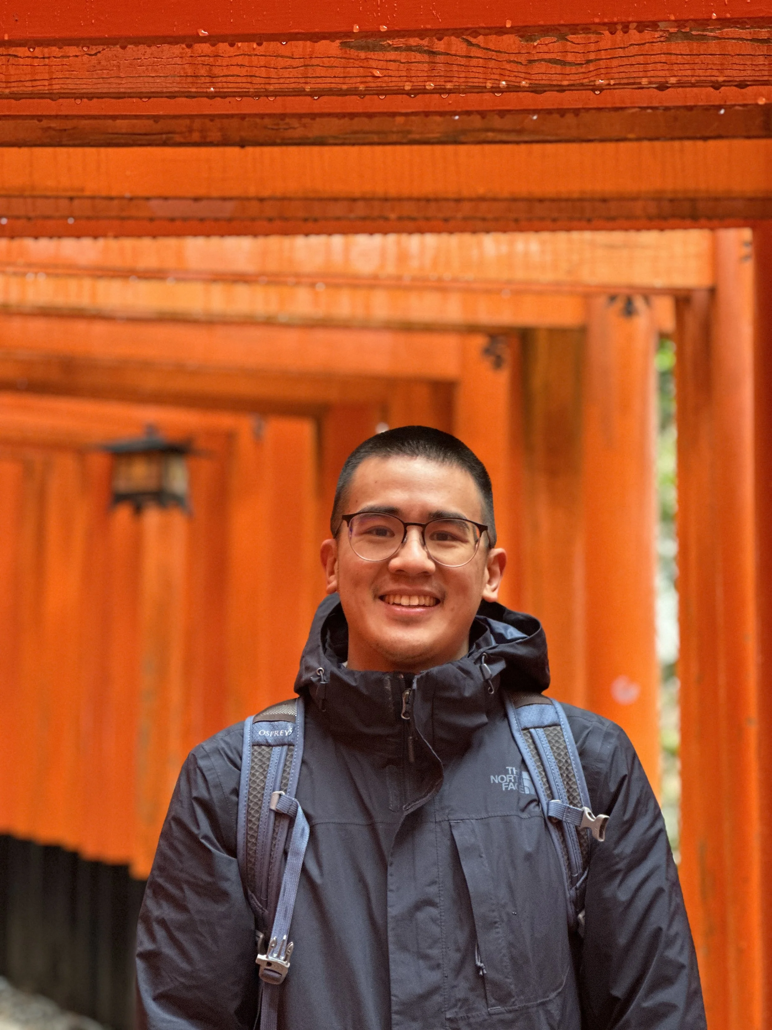 A smiling man with glasses, wearing a black North Face jacket and a backpack, standing in front of a series of red torii gates at a Japanese shrine.