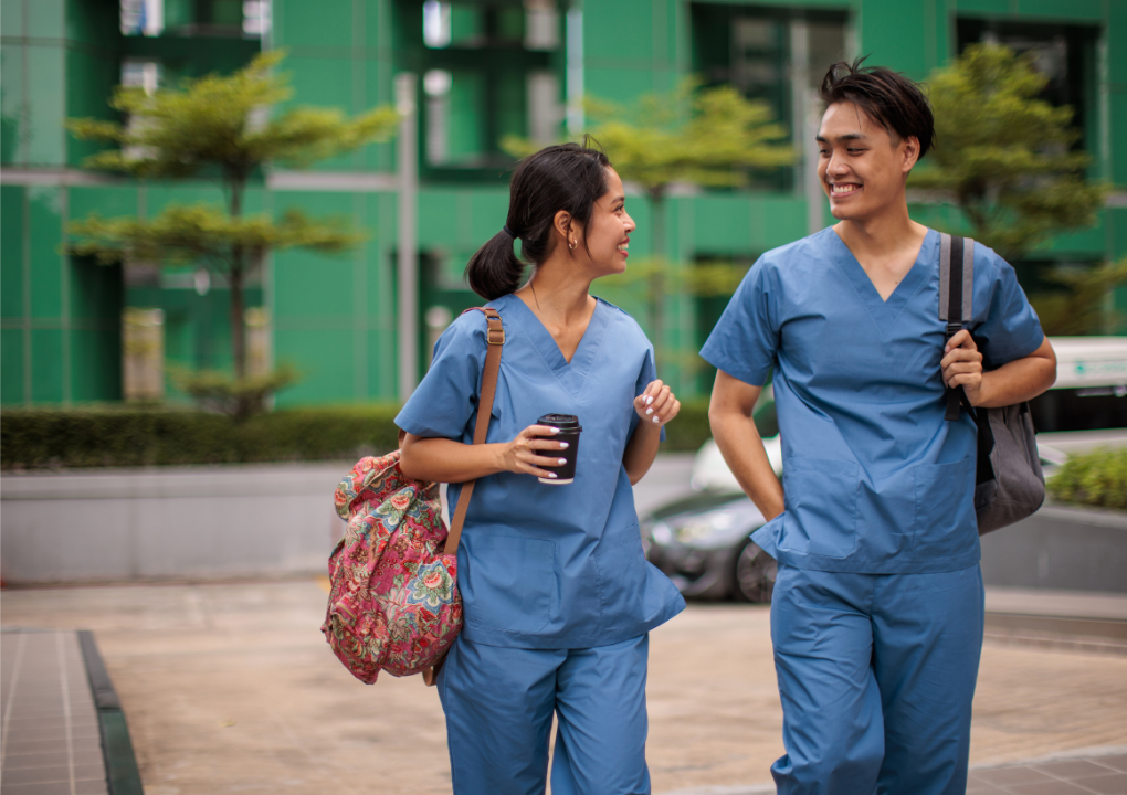 Two healthcare professionals dressed in blue scrubs walking and talking outside, smiling at each other, carrying bags and coffee cups.