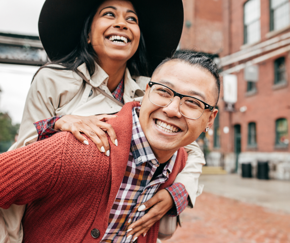 Two friends laughing and smiling outdoors on a city street, with red brick buildings in the background. One person is giving the other a piggyback ride, both appear joyful.