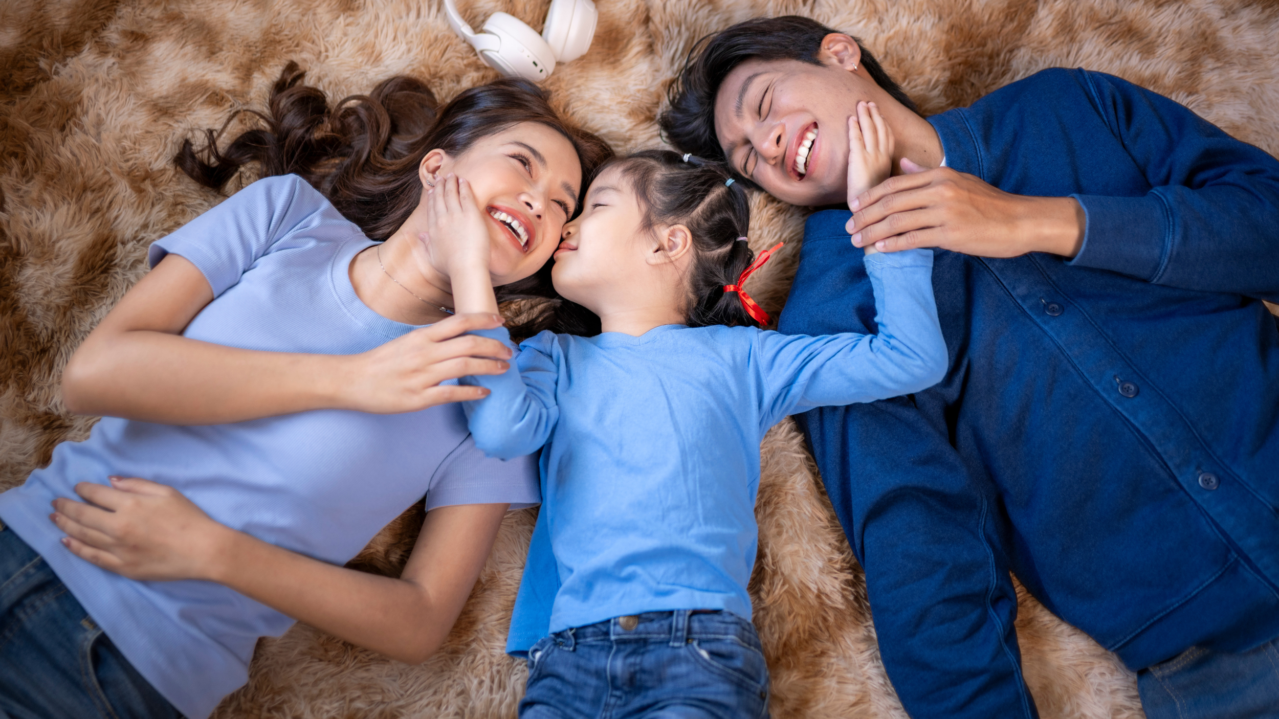 A family of three lying on a fluffy brown rug, smiling and enjoying each other's company, with a white pair of headphones nearby.