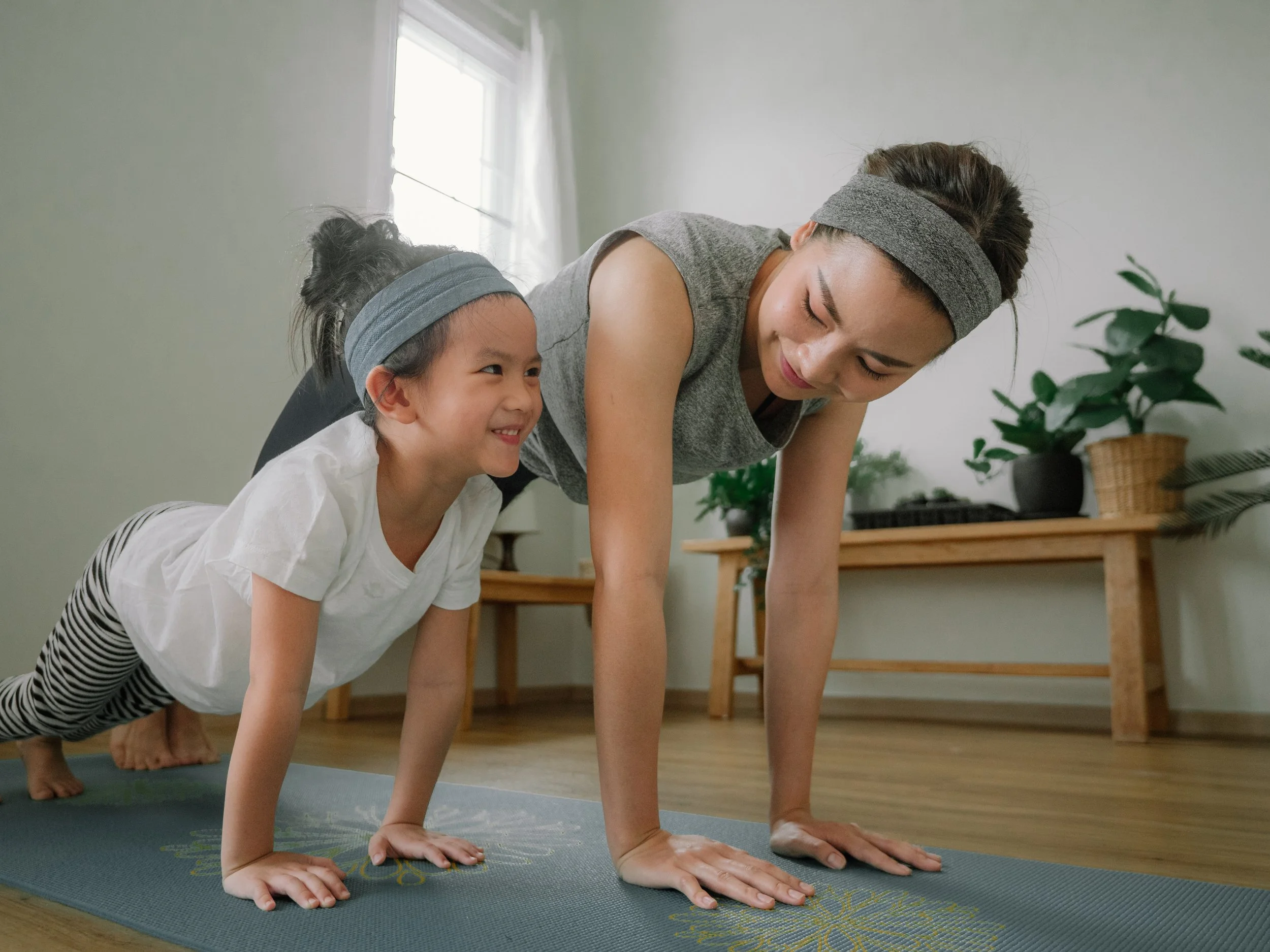A woman and a young girl in workout clothes practicing push-ups on a yoga mat indoors, with greenery in the background.