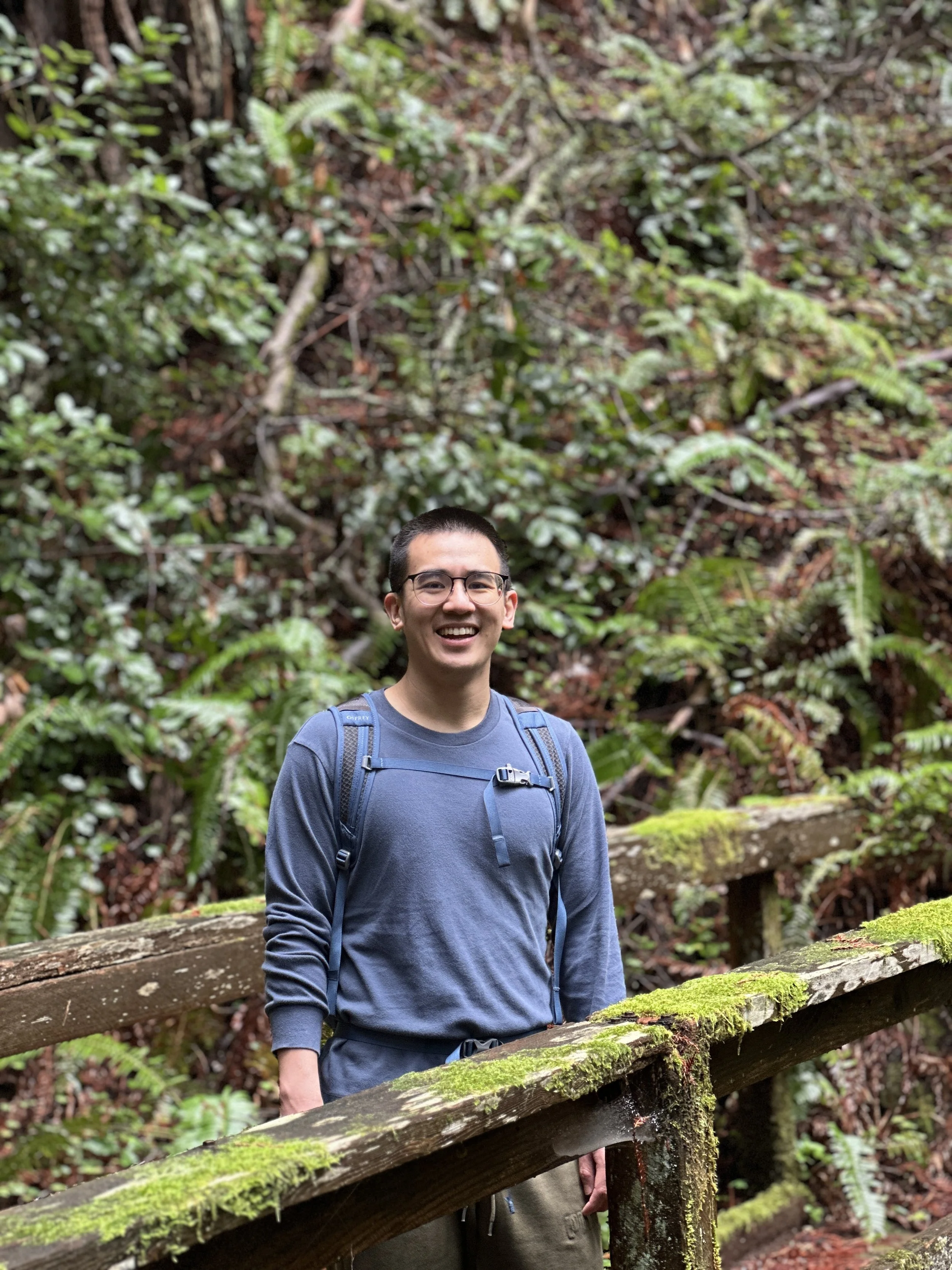A man with glasses and a backpack smiling, standing on a mossy wooden bridge in a lush, green forest.