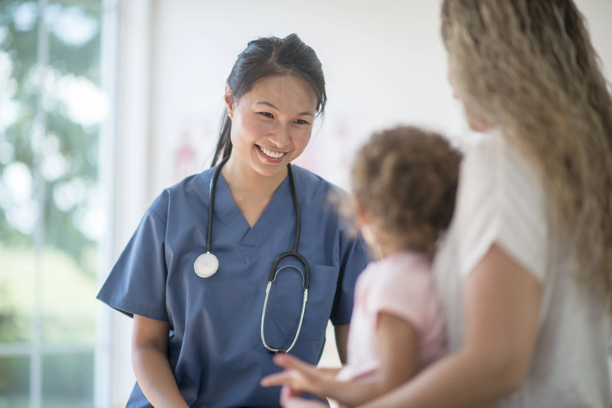 A female nurse in blue scrubs smiling and talking to a woman with two children in a bright medical facility.