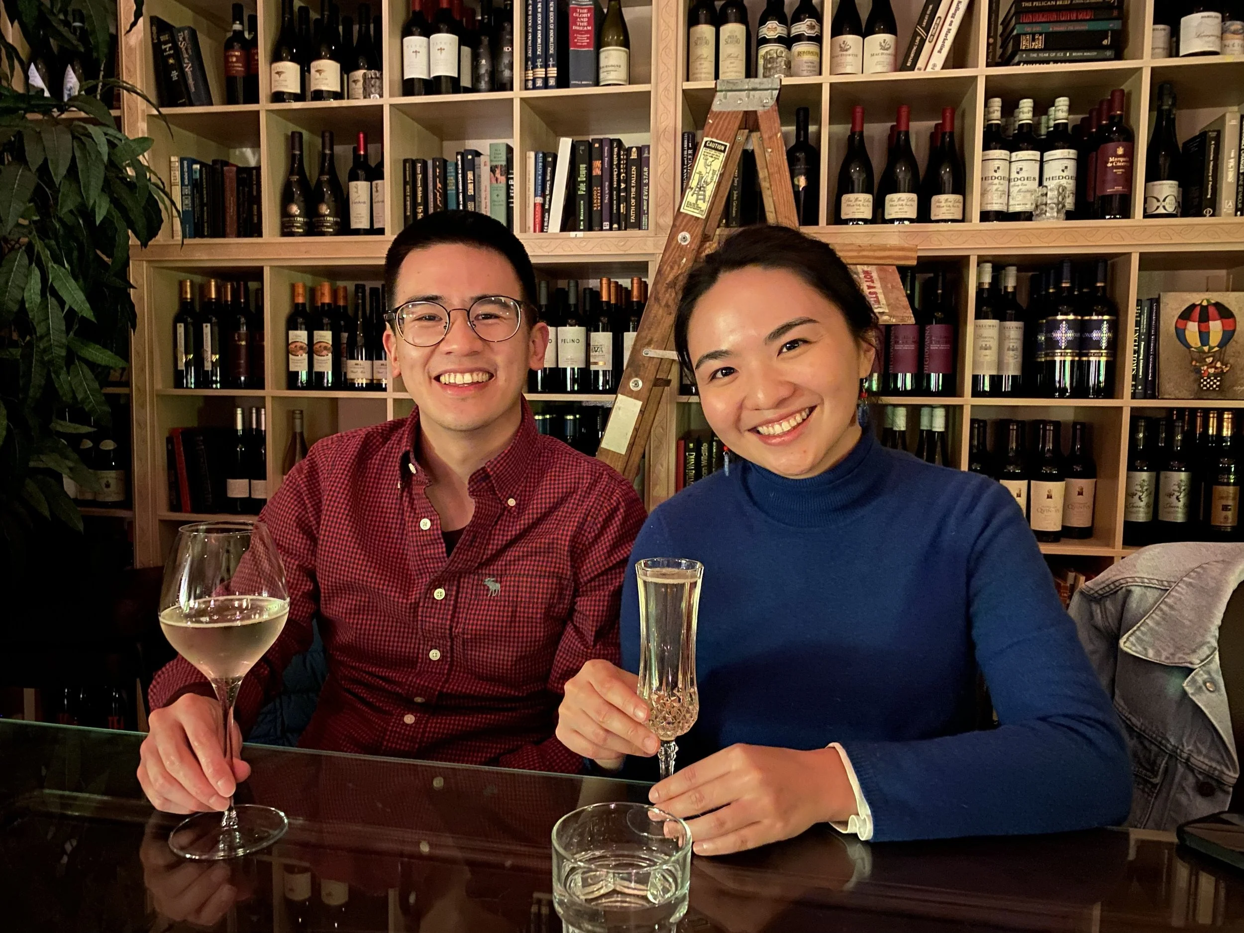 Two smiling people, a man wearing glasses and a red checkered shirt and a woman wearing a blue sweater, sitting at a table in a room with wooden shelves stocked with wine bottles and books, holding wine glasses.