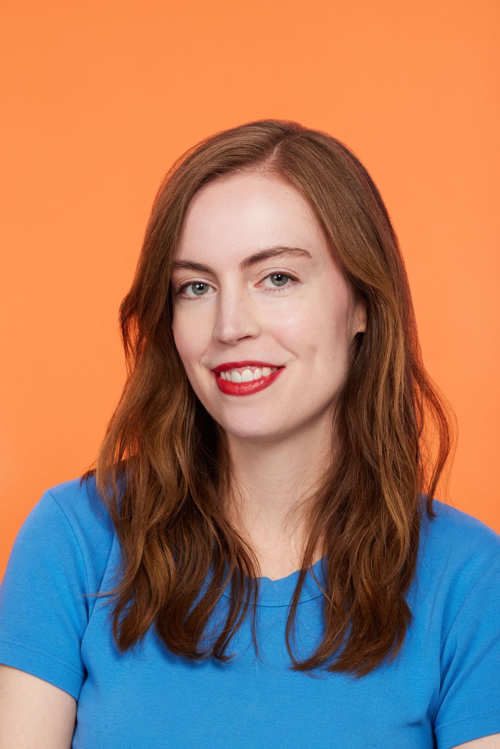 A woman with long, wavy reddish-brown hair, blue eyes, and red lipstick, smiling at the camera in front of an orange background, wearing a blue shirt.