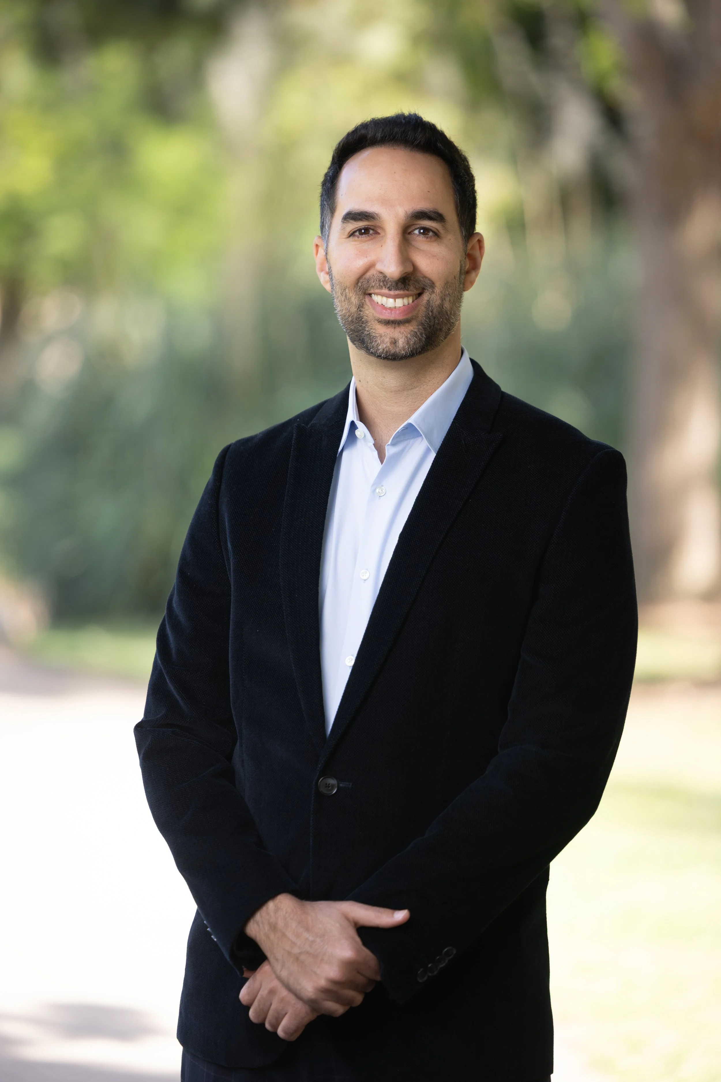 A man wearing a black blazer and white shirt standing outdoors with a blurred nature background.