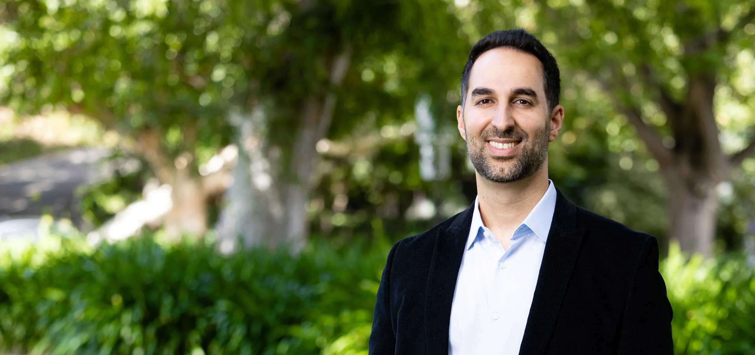 A smiling man with dark hair and a beard is standing outdoors in front of green trees and foliage, wearing a dark blazer and a light blue shirt.
