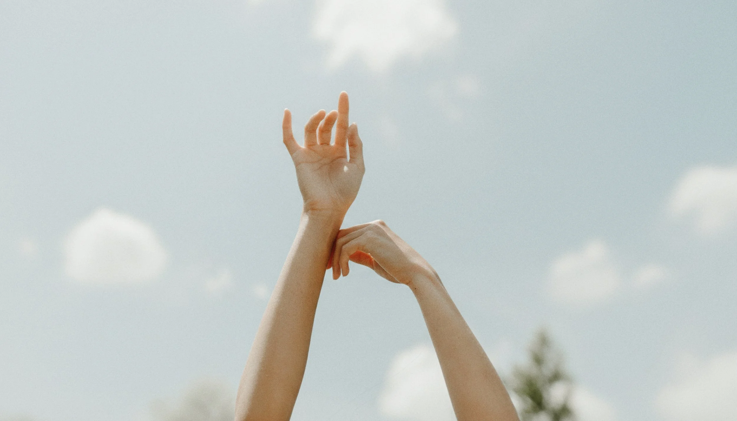 Two hands with fingers intertwined and one hand's index finger pointing upwards against a cloudy sky.