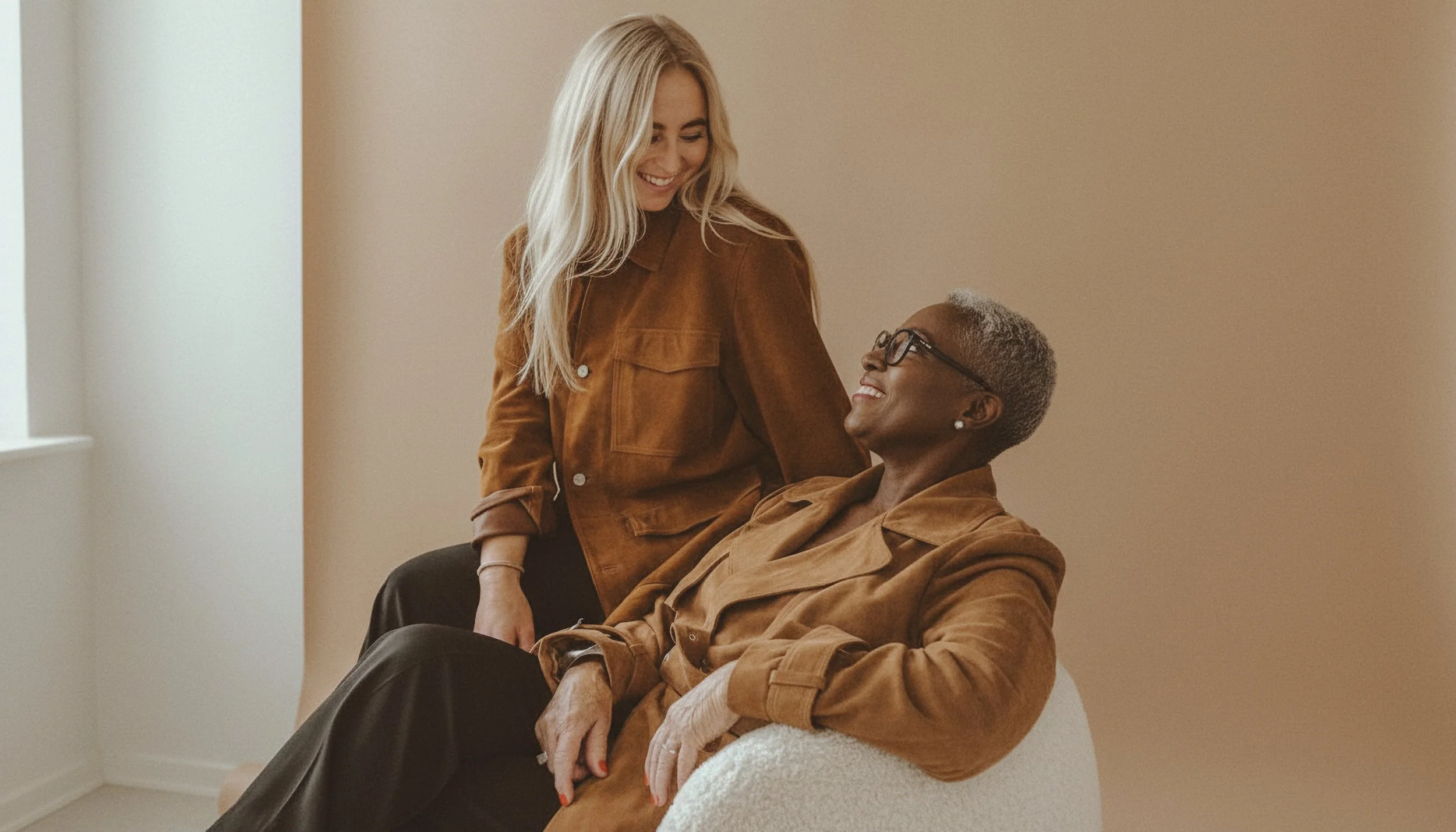 A young woman with long blond hair smiling at an older woman with short gray hair and glasses, sitting on a chair, both wearing brown jackets, in a neutral-colored room.