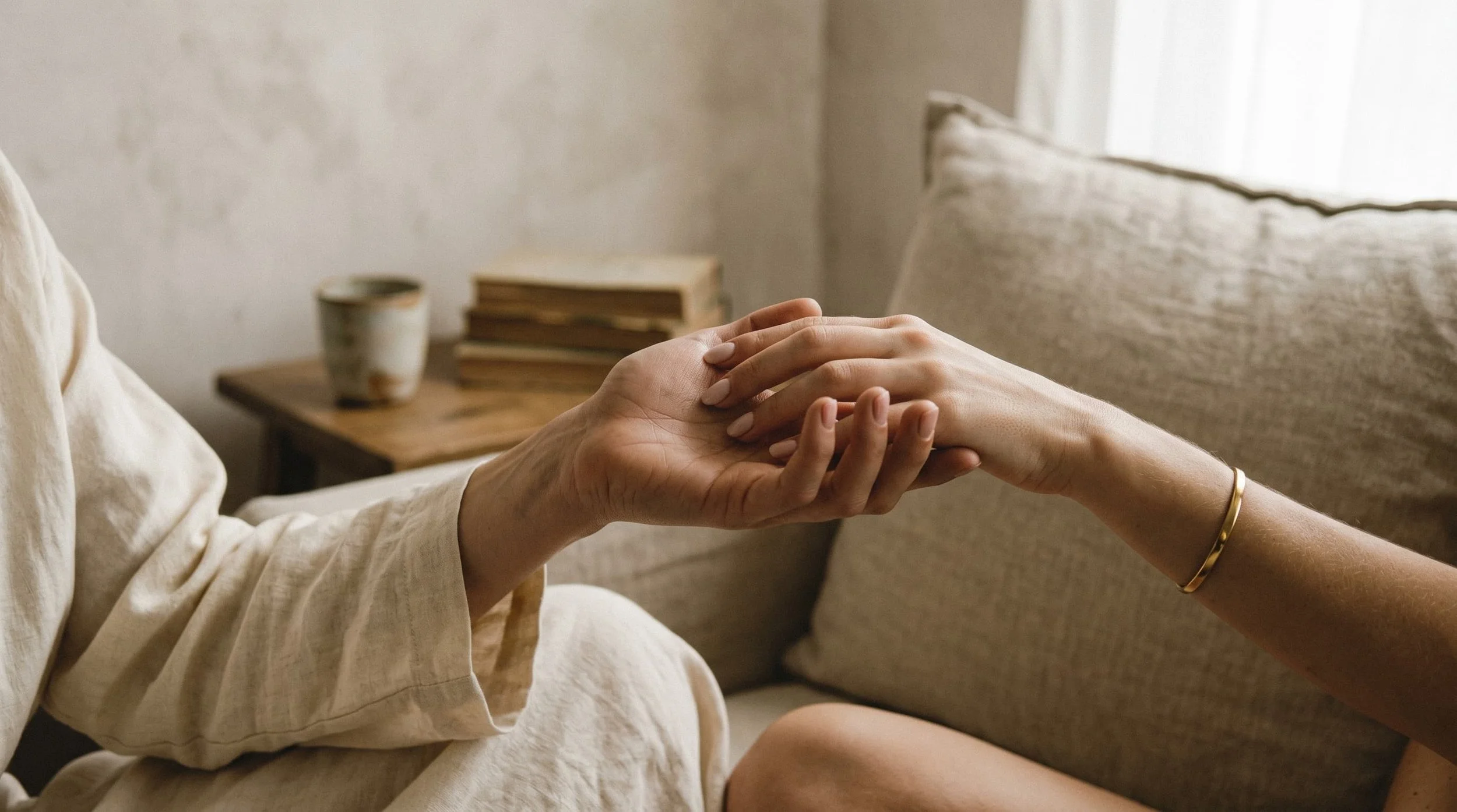 Two people holding hands, with a woman wearing a gold bracelet, in a cozy room with a beige couch and a wooden side table with cups and books.