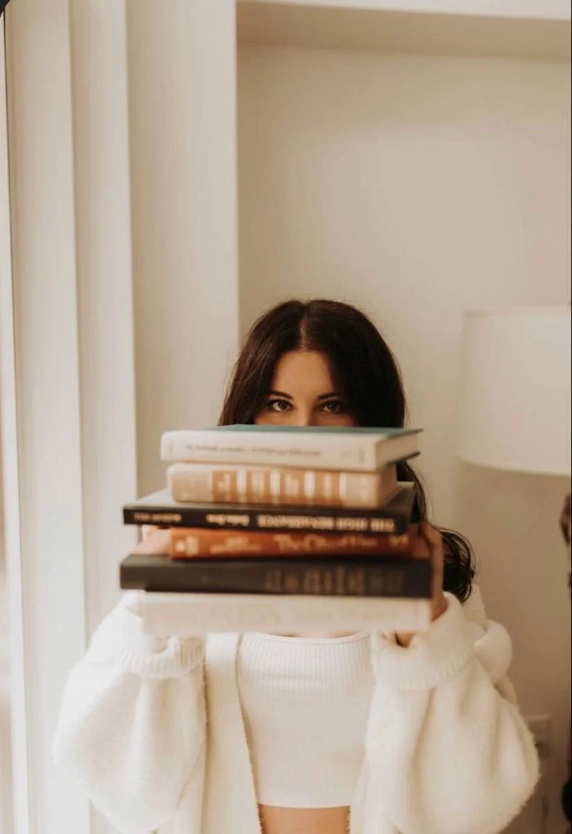 A woman with dark hair holding a stack of books in front of her face, partially hiding her face, in a cozy interior setting.