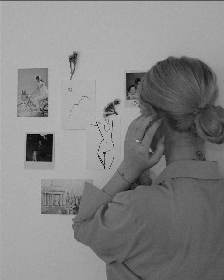 A woman with blonde hair tied in a bun, wearing a light-colored shirt, is standing with her back to the camera, looking at a wall with various photos and drawings. She is holding a phone to her ear with her right hand.