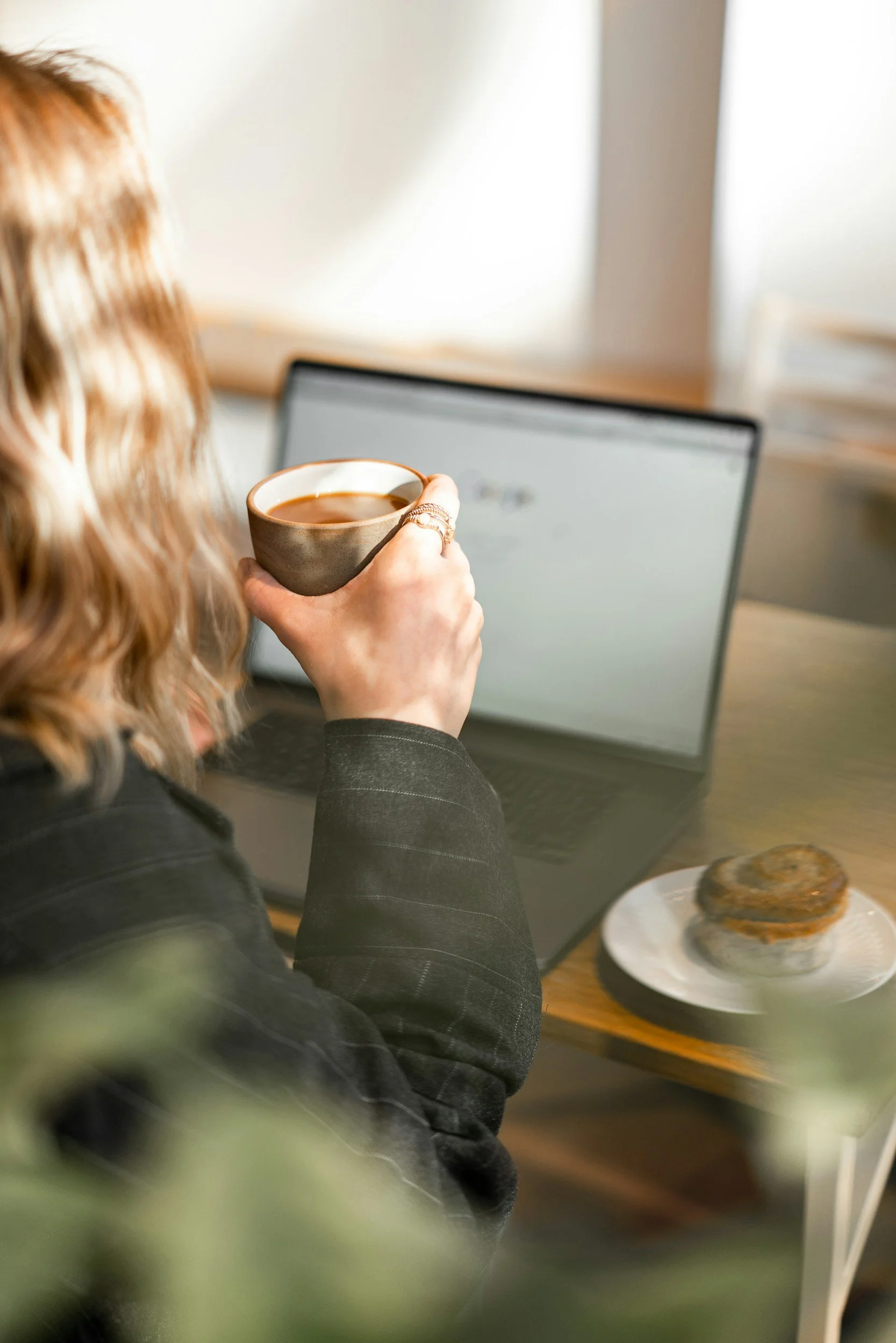 A woman with blond wavy hair wearing a dark blazer, holding a cup of coffee, sitting at a desk with a laptop and a plate of muffins.
