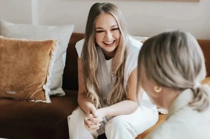 Two women sitting on a couch, smiling and laughing while talking to each other in a cozy living room.