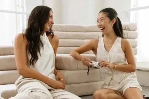 Two women sitting on a sofa, smiling and chatting, with one woman holding a camera or phone.