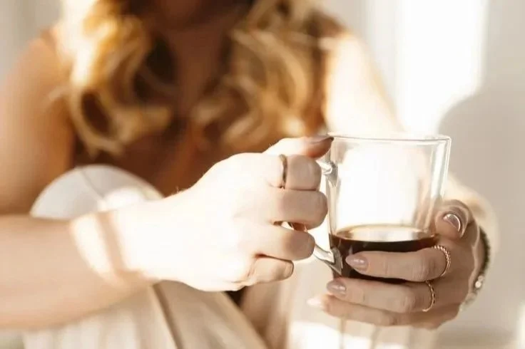 A woman with long, wavy hair is holding a transparent glass mug with a dark beverage, likely coffee or tea.