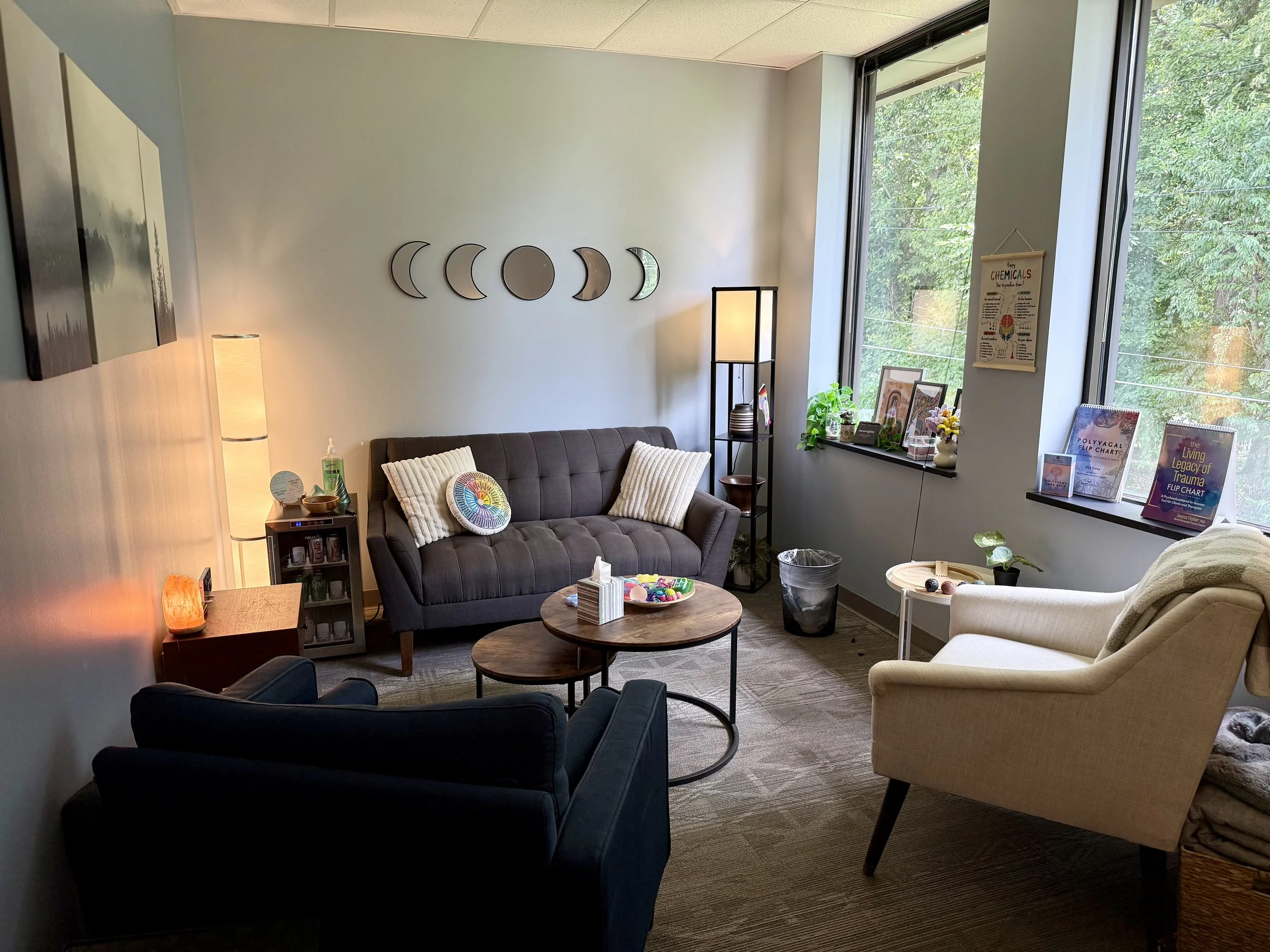 A cozy waiting room with a brown sofa, a beige armchair, a small round wooden coffee table, and decor including a moon phase wall art, books, plants, and framed pictures near large windows with a view of green trees outside.