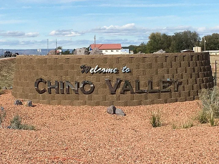 Welcome sign for Chino Valley on a brown brick wall with rocks and desert plants in front, under a partly cloudy sky.