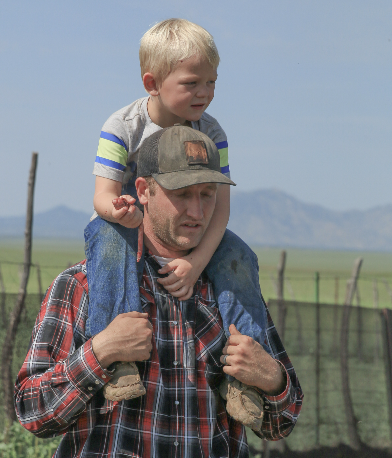 A man carrying a young boy on his shoulders outdoors in a rural area, with mountains in the background and a fence in the foreground.