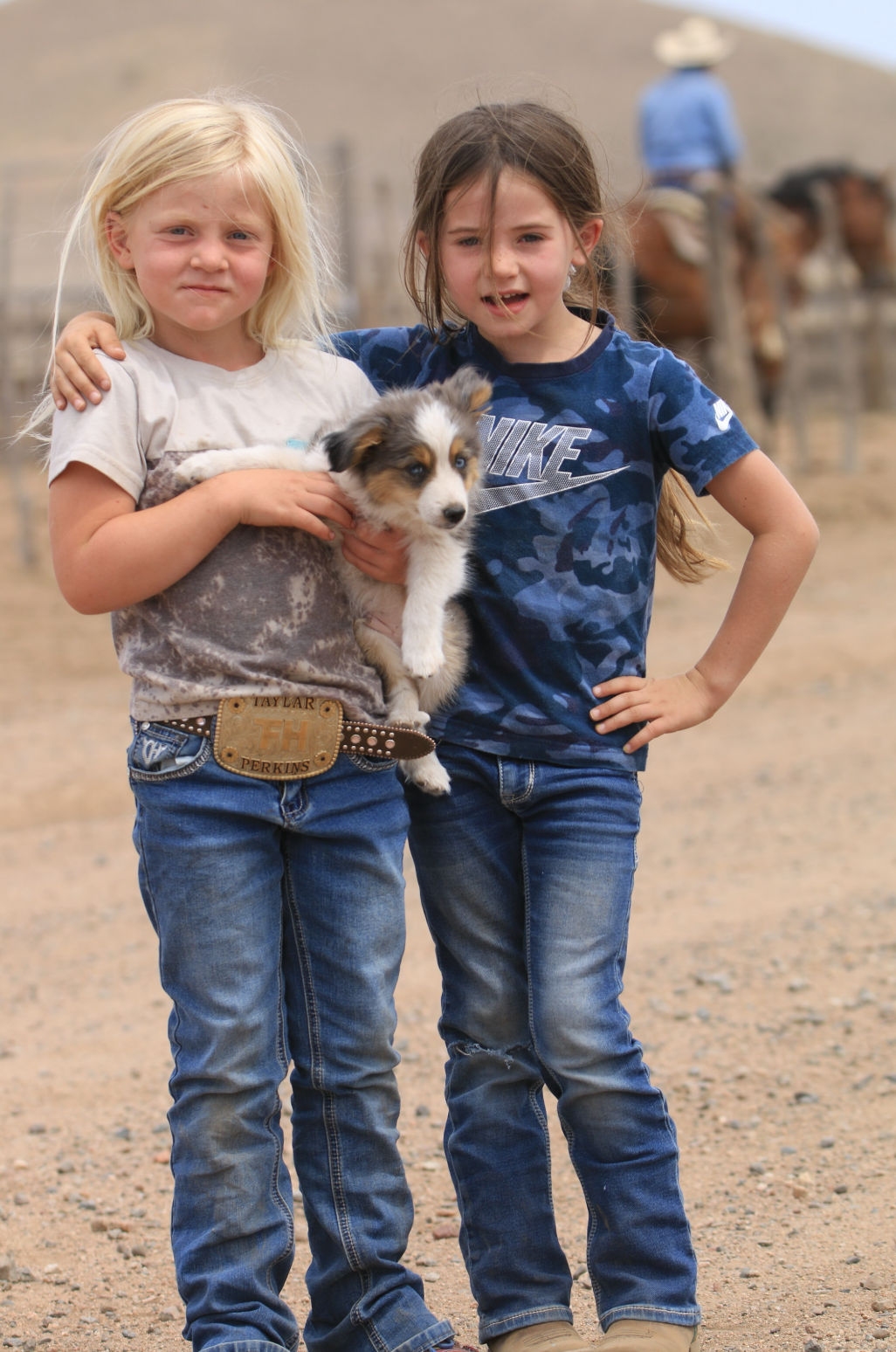 Two young girls are standing outdoors on a dirt path, with one girl holding a puppy. The girl on the left has blonde hair, is wearing a beige t-shirt and jeans with a belt. The girl on the right has brown hair in a ponytail, is wearing a blue camouflage t-shirt and jeans. There are horses and a person in the background.
