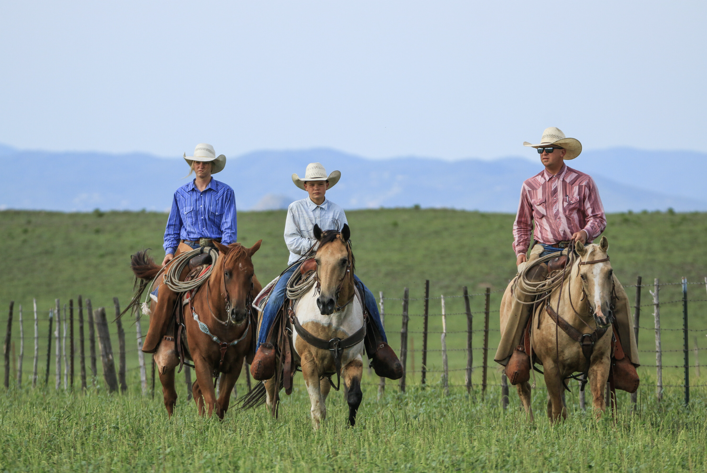 Three people riding horses across a grassy field, wearing cowboy hats and western attire, with a mountain range in the background.