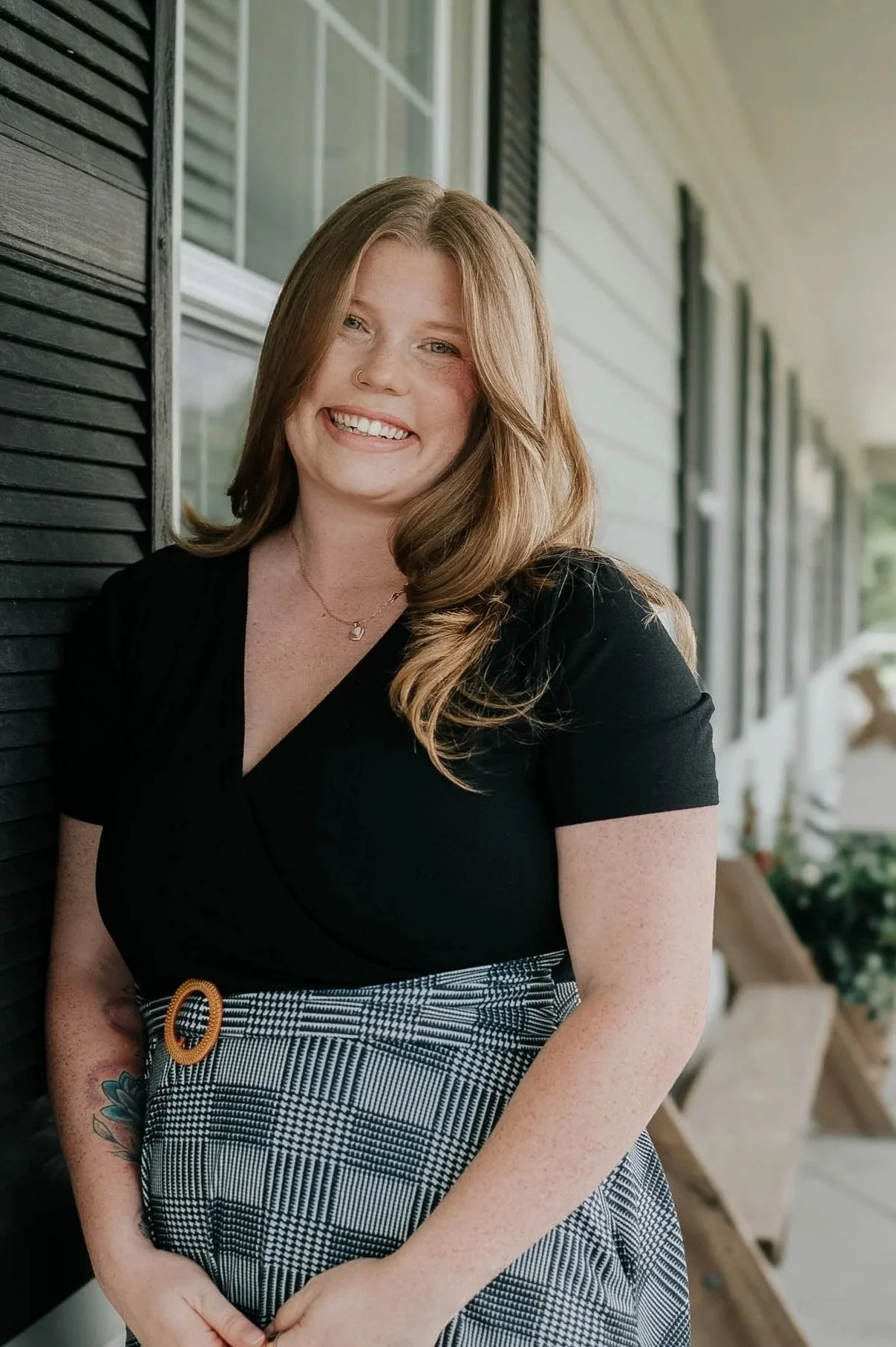 A woman with long, wavy red hair smiling, wearing a black top and plaid skirt, standing outdoors with a porch and windows in the background.