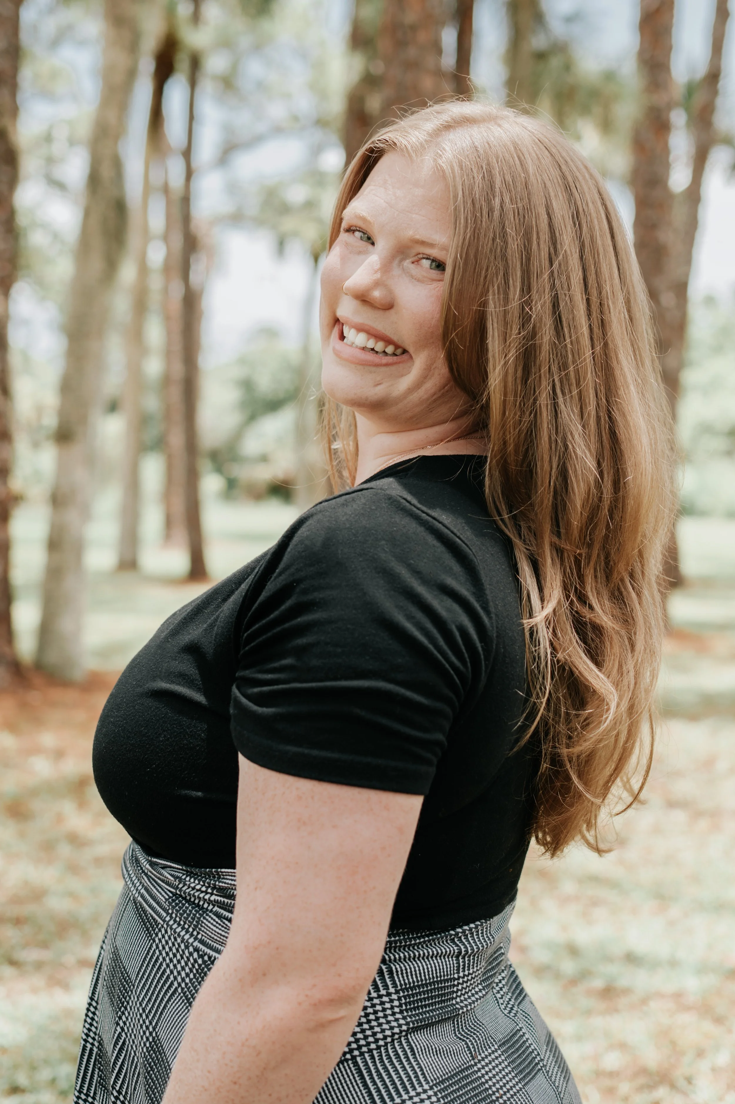 A young woman with long red hair smiling outdoors, standing near trees, wearing a black top and plaid skirt.