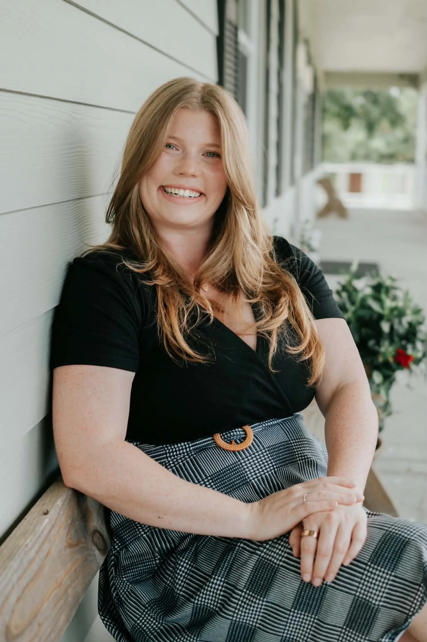 A woman with long wavy reddish-brown hair and a nose ring sitting on a porch beside a white wall, smiling and looking at the camera. She is wearing a black top and a checkered skirt with a brown buckle belt, with her arms crossed over her lap.