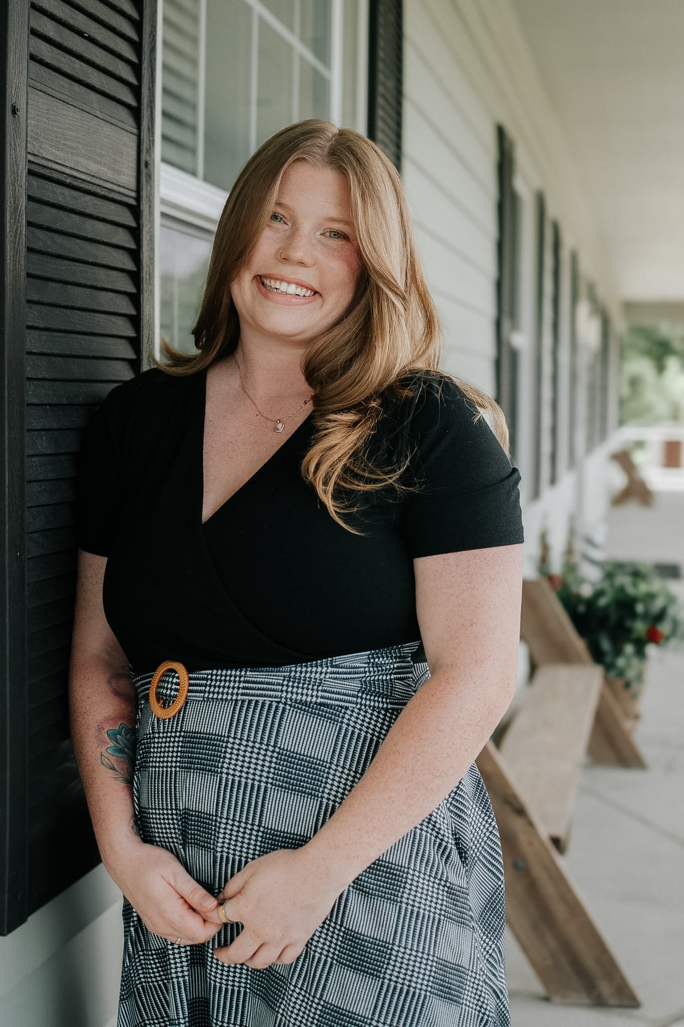 A woman with long red hair smiling and leaning against a house exterior wall with black shutters behind her.