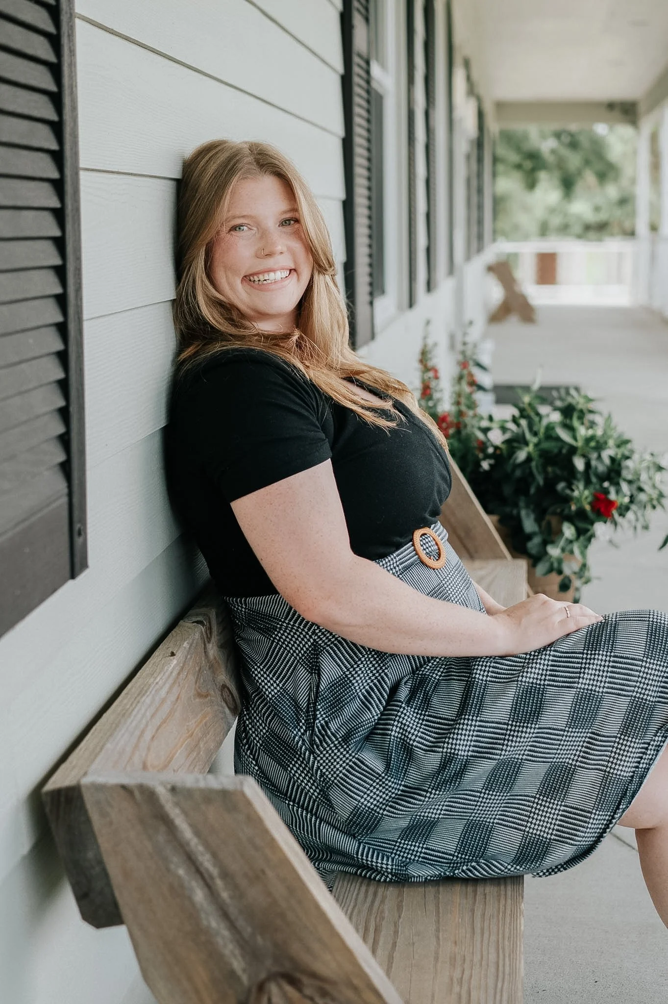 A smiling woman with blonde hair, wearing a black top and patterned skirt, sitting on a wooden bench on a porch with a light gray wall and potted plants in the background.