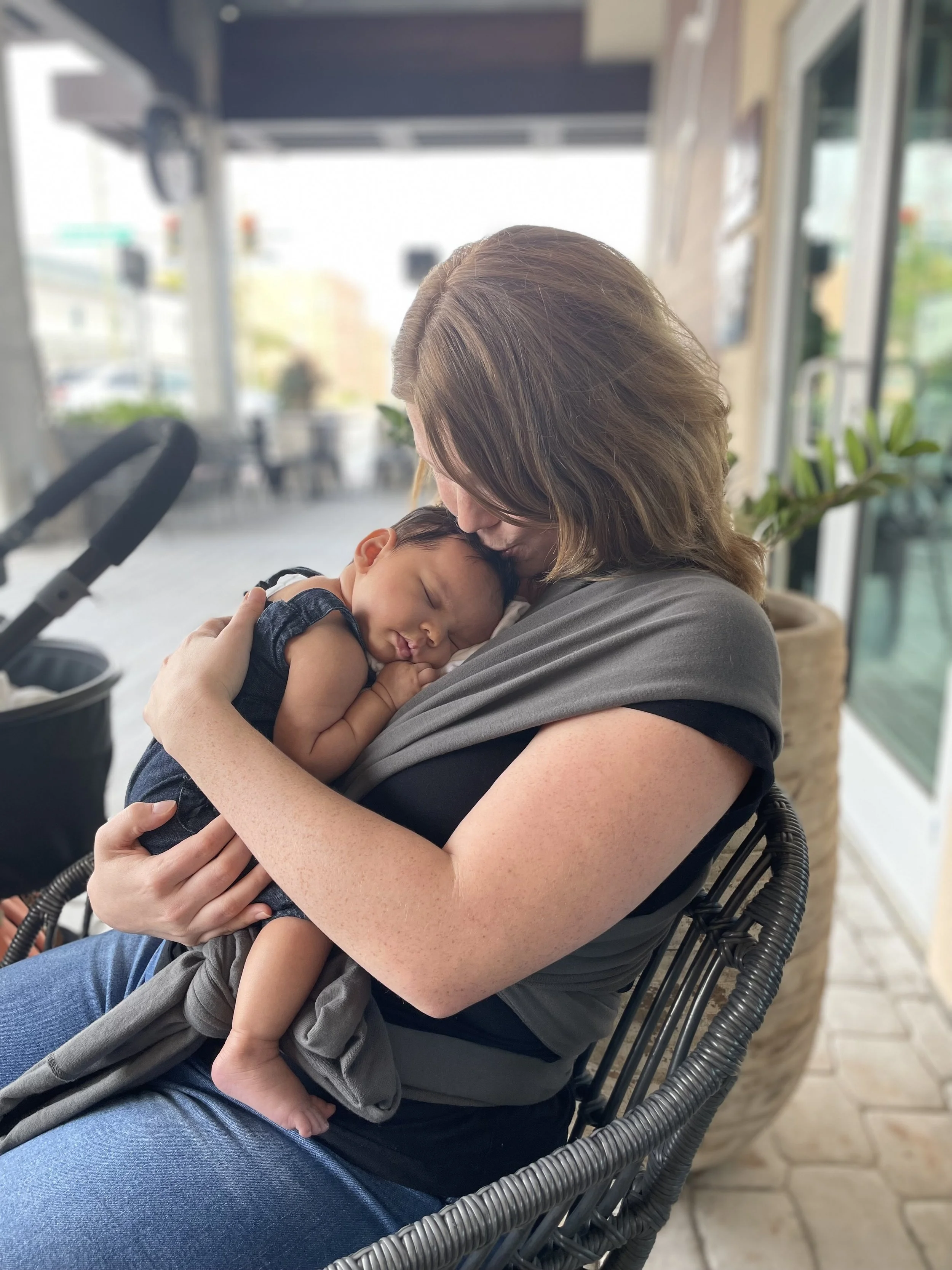 A woman with brown hair is holding a sleeping baby with short dark hair and closed eyes, with her face kissing the baby's forehead in an indoor setting with glass walls and plants.