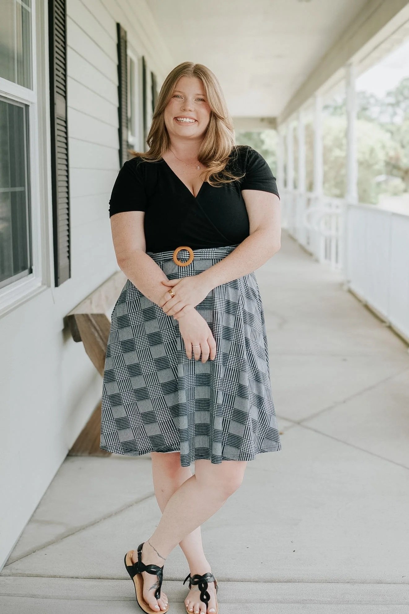 A woman standing outside on the porch of a house, smiling at the camera, wearing a black top and a plaid skirt with a belt, and black sandals.