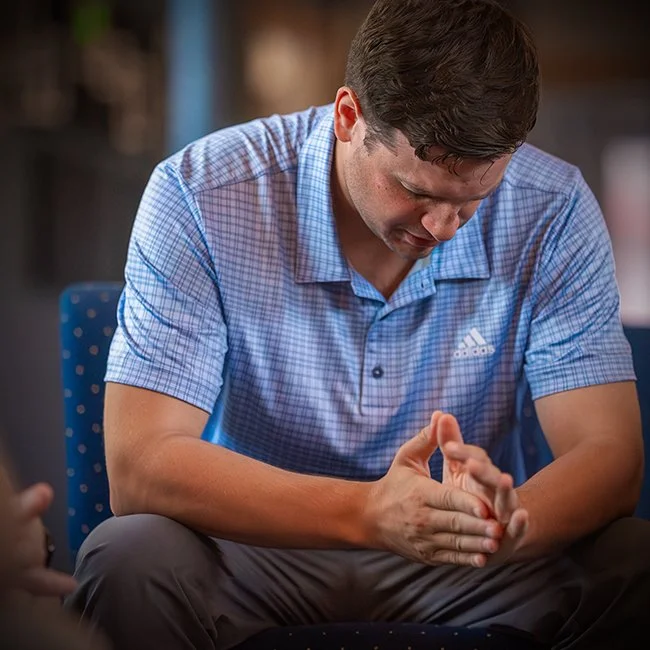A man in a blue checkered shirt sitting down, with his hands clasped and head bowed in a prayer or moment of reflection.