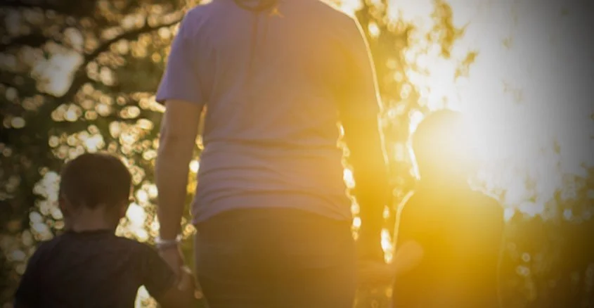 A woman holding hands with two children outdoors during sunset, with trees in the background.